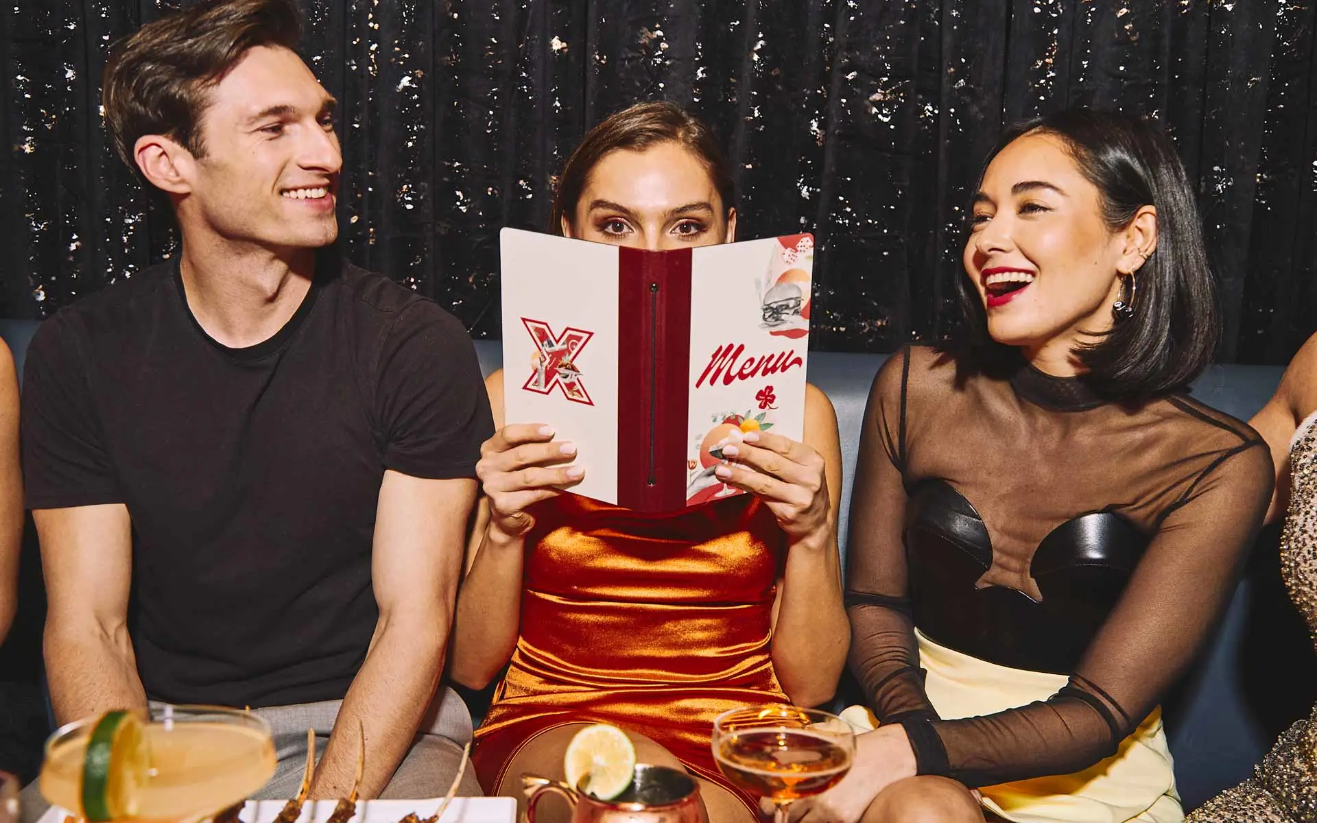 Three young adults seated in a stylish lounge booth, laughing and socializing while one person holds a Lucky Strike menu.