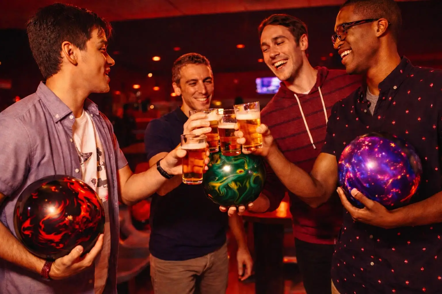 A group of four friends standing near bowling balls and raising glasses in a cheerful toast inside a bowling center.