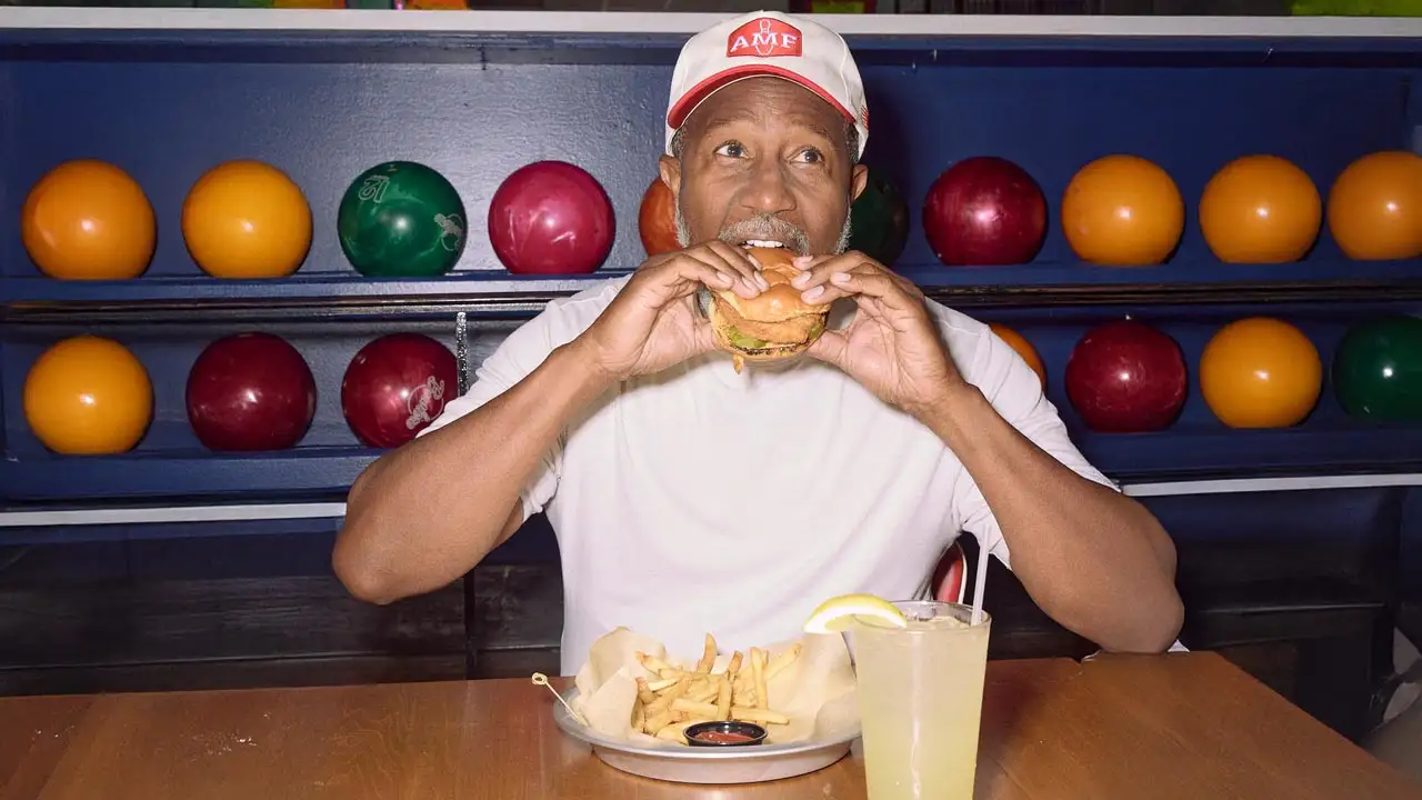 Man eating a chicken sandwich with fries and a lemonade in front of a rack of bowling balls at an AMF bowling center.