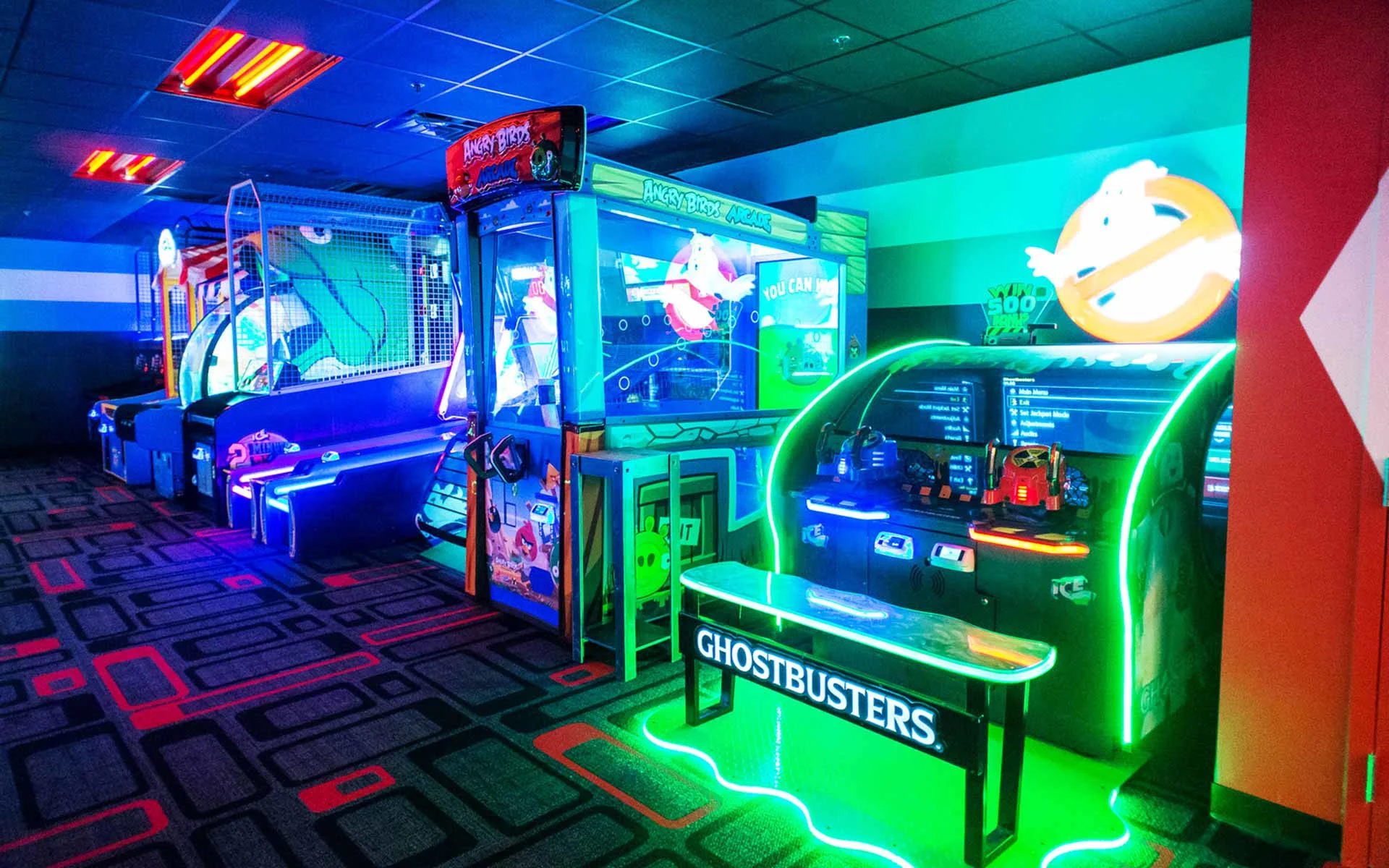 Vibrant arcade interior featuring neon-lit game machines, including a Ghostbusters arcade cabinet in the foreground, alongside air hockey and other interactive games, set against colorful lighting and patterned carpet in a modern entertainment venue.