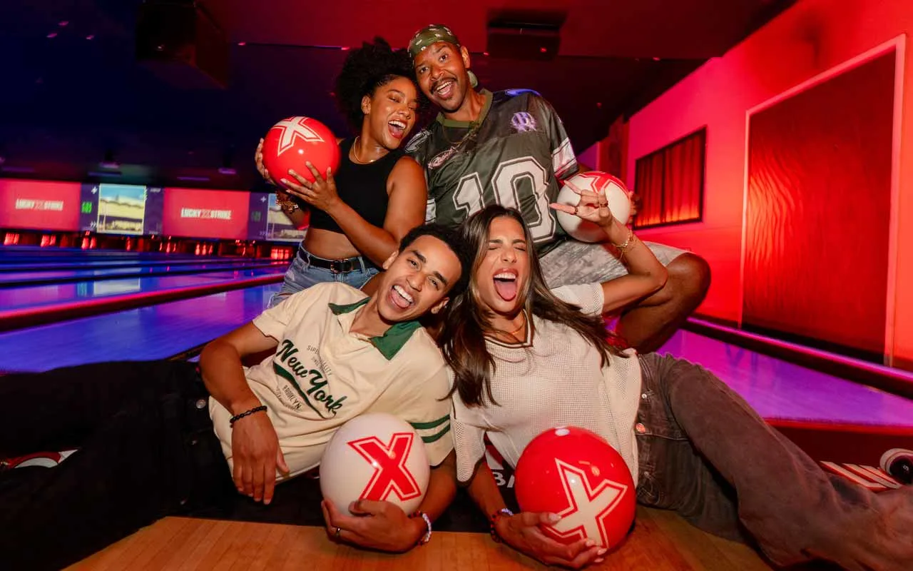 Group of friends laughing and posing with Lucky Strike branded bowling balls on the lanes under colorful neon lighting.