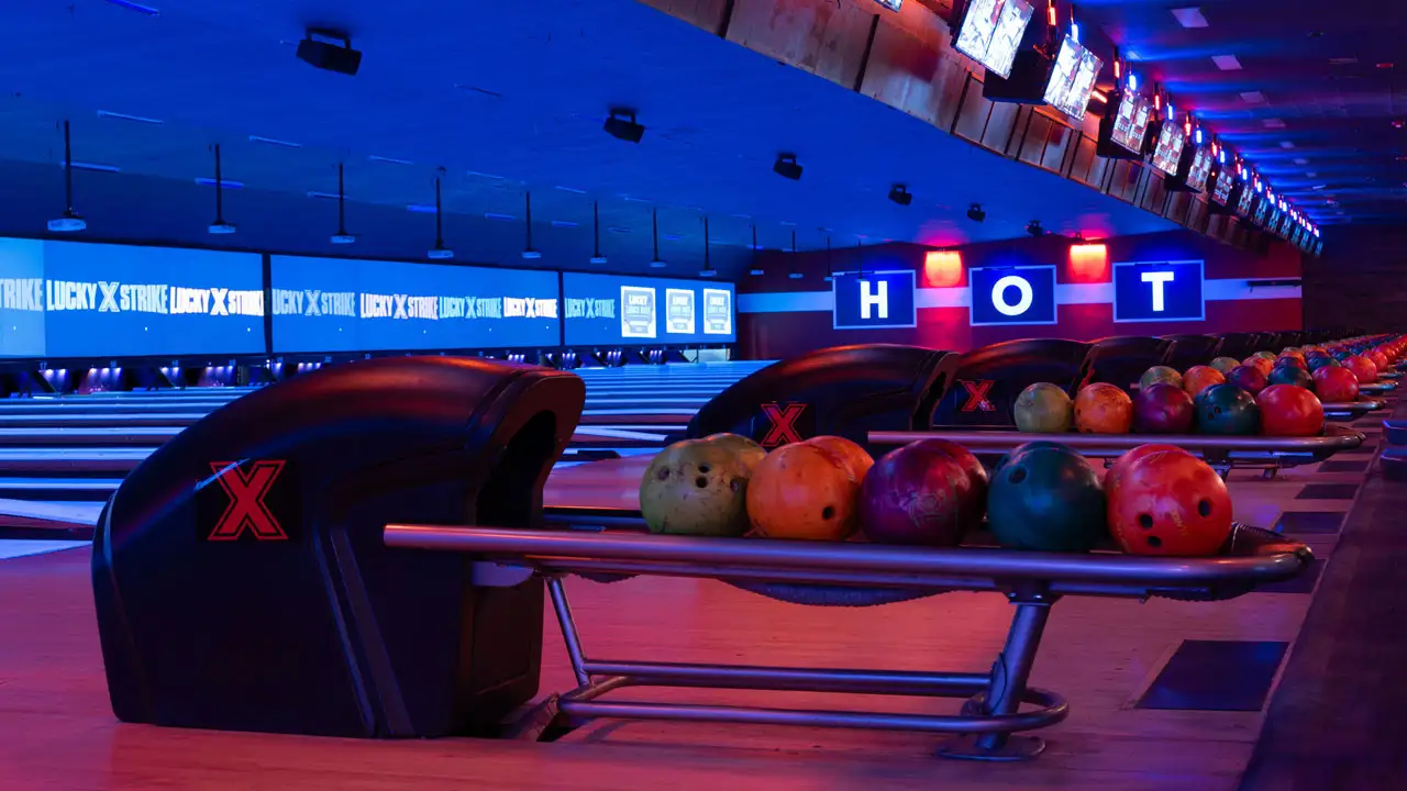 Colorful bowling balls in a return rack with bowling lanes and large TV screens in the background.