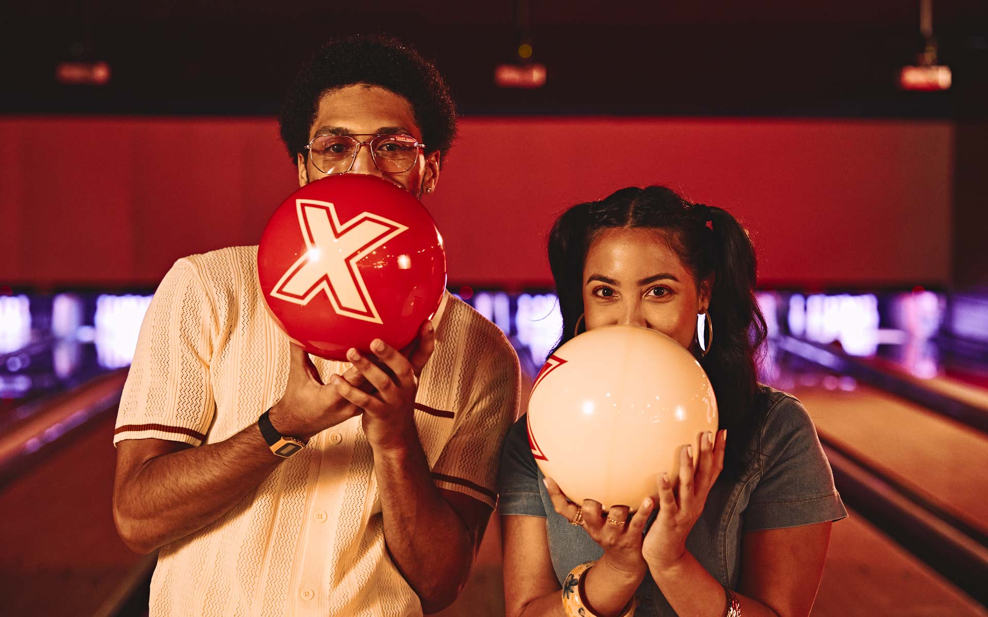 Couple holding bowling balls over their faces in a warmly lit bowling alley, playful pose on lane with red and cream balls.