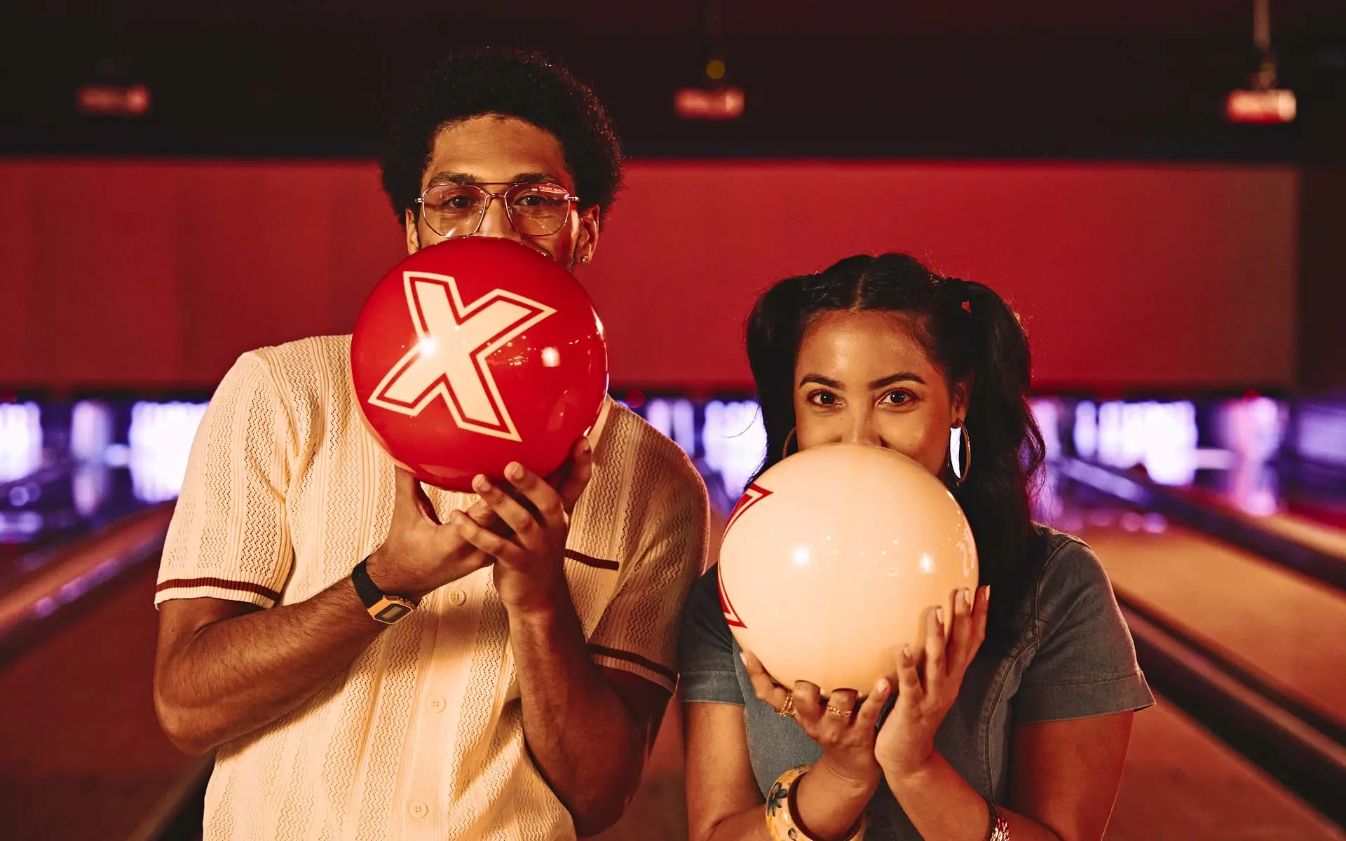 Couple holding bowling balls over their faces in a warmly lit bowling alley, playful pose on lane with red and cream balls.