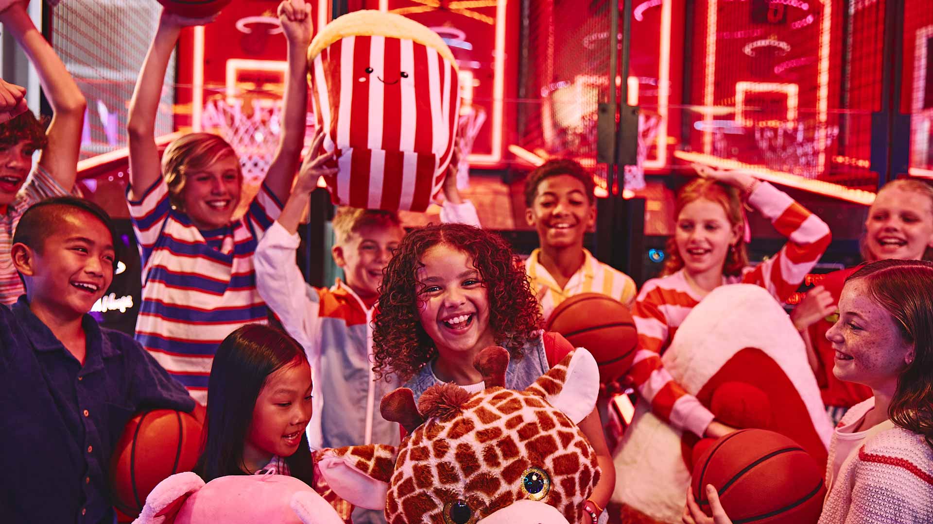 Children smiling and holding basketballs and plush prizes while playing arcade games in a brightly lit entertainment center.