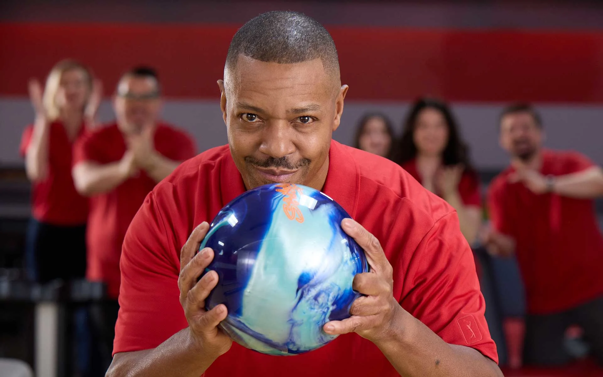 Man in red shirt aiming a blue bowling ball at an AMF bowling alley with teammates cheering in the background