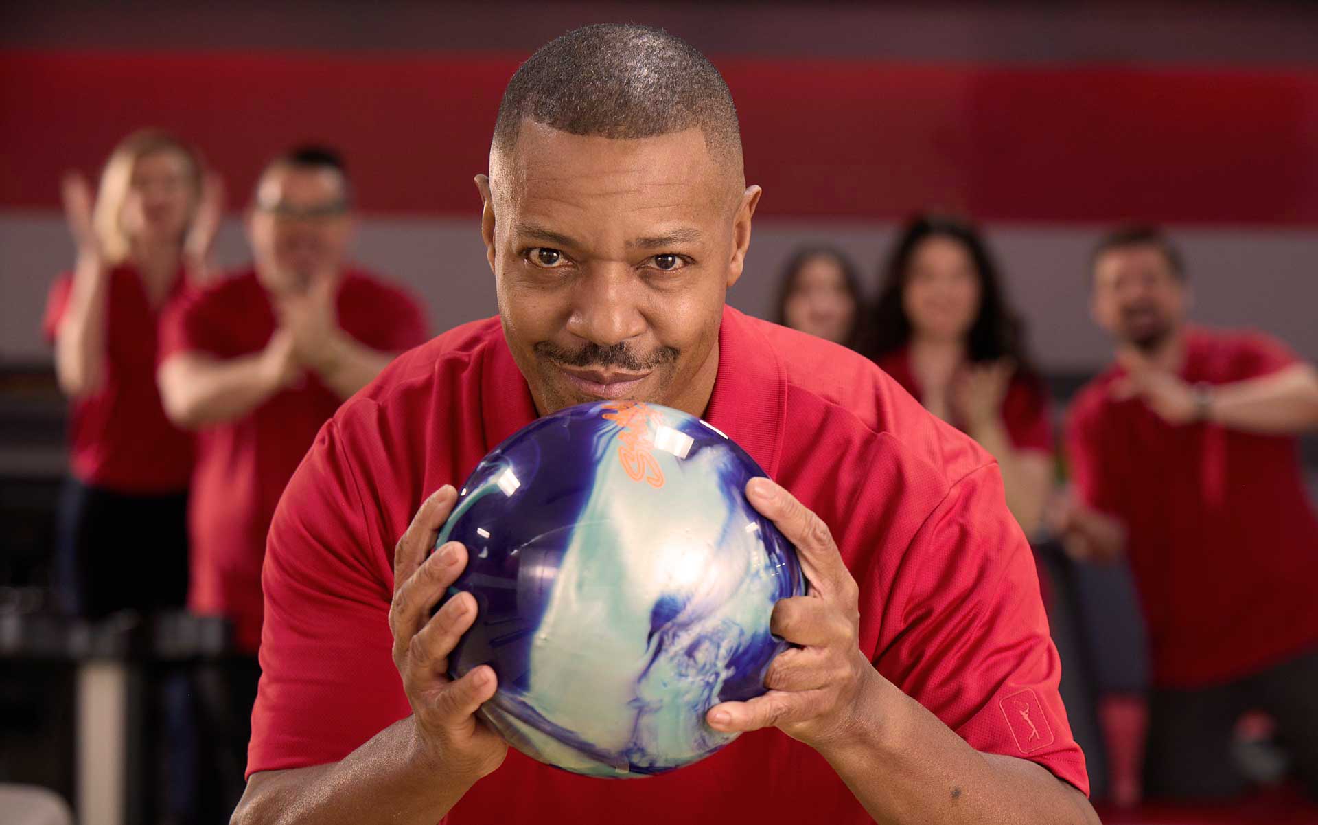 Man in red shirt aiming a blue bowling ball at an AMF bowling alley with teammates cheering in the background.