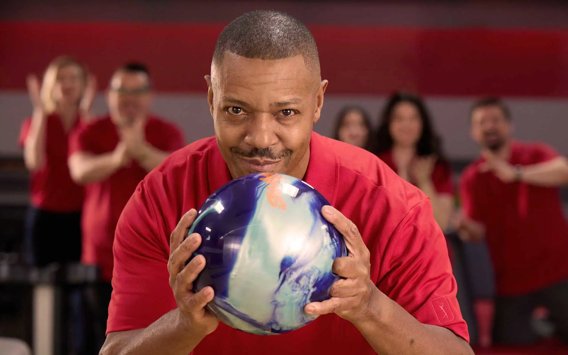Man in red shirt aiming a blue bowling ball at an AMF bowling alley with teammates cheering in the background.