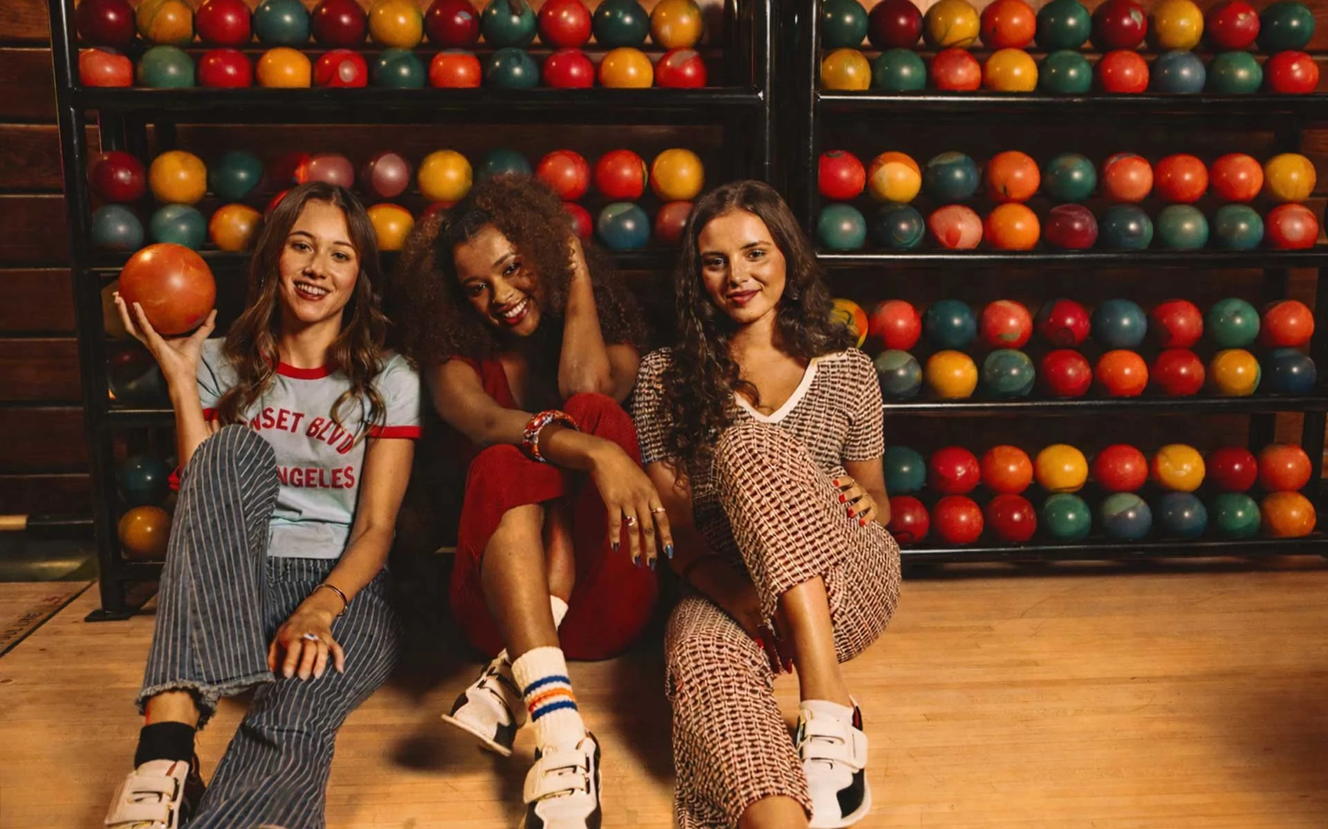 Three young women sitting on a bowling alley floor in front of a rack filled with colorful duckpin bowling balls, smiling at the camera, with one holding a small orange duckpin ball under warm, ambient lighting.