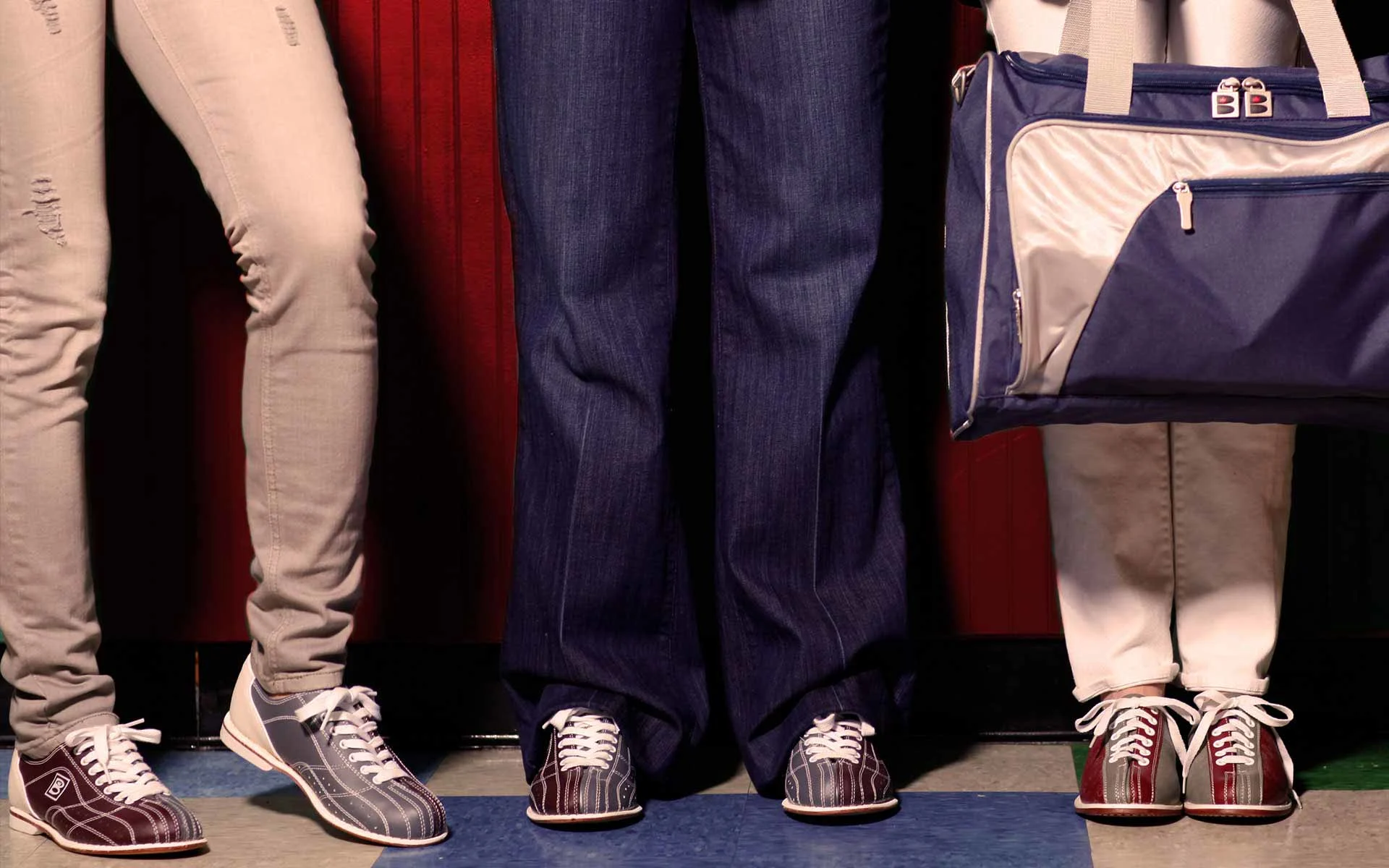 Three people standing side by side wearing bowling shoes; only their legs and feet are visible. One person holds a blue and white bowling bag.