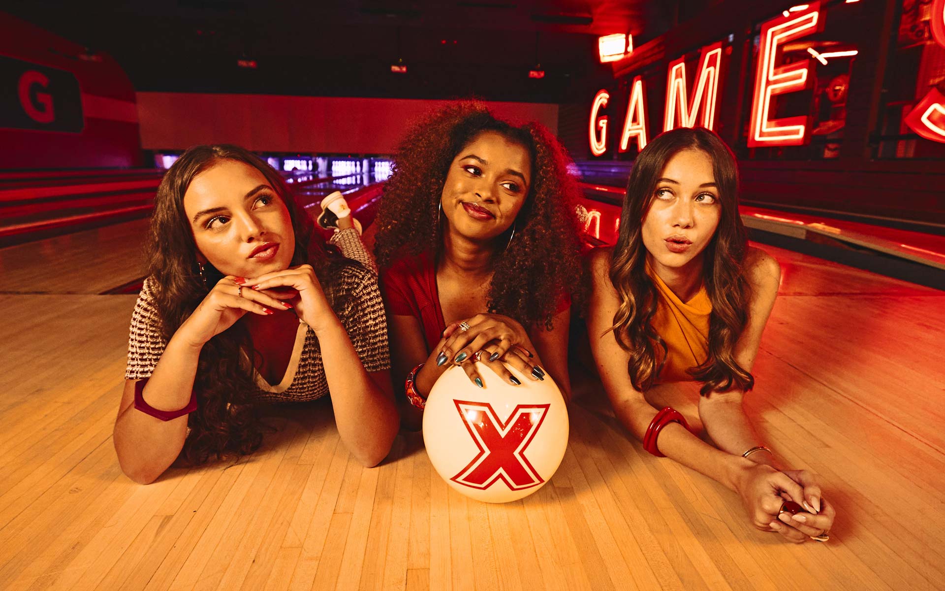 Three women pose on a bowling lane in a neon-lit bowling alley with a white bowling ball, glowing red signage, and illuminated pins in the background.