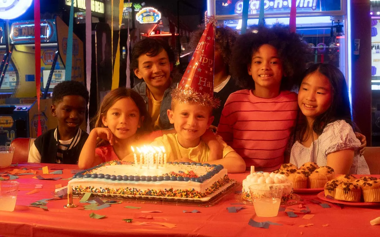 Group of children gathered around a birthday cake with lit candles at an arcade party, smiling and celebrating among colorful decorations, cupcakes, and glowing game machines in the background.