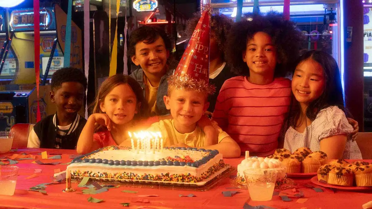 Group of children gathered around a birthday cake with lit candles at an arcade party, smiling and celebrating among colorful decorations, cupcakes, and glowing game machines in the background.