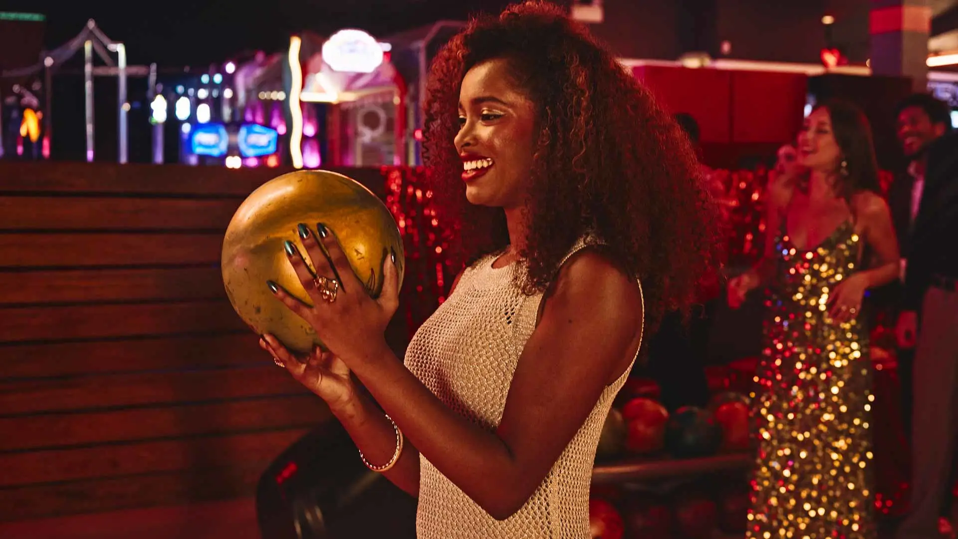 Woman smiling and holding a bowling ball at an upscale bowling venue while guests socialize in the background.