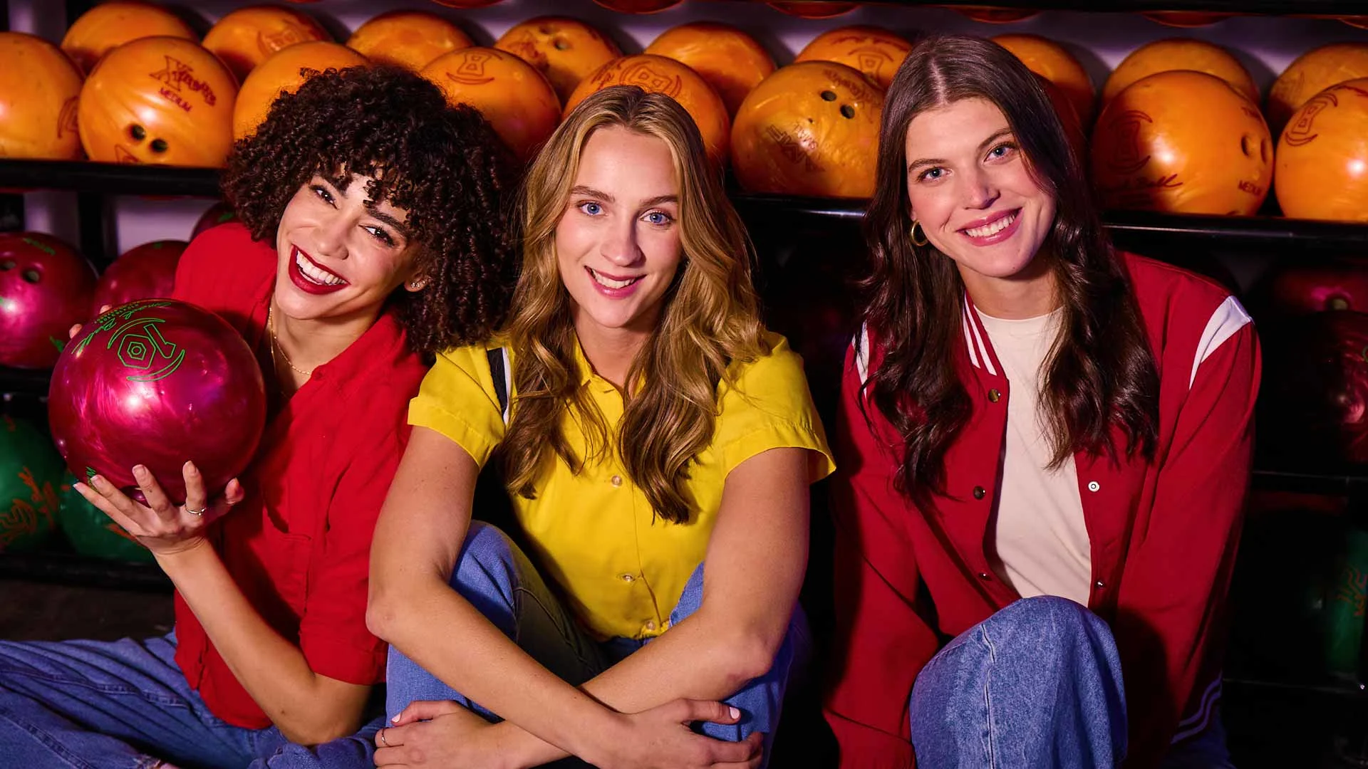 Three women sit smiling in front of a bowling ball rack, with one holding a red bowling ball and rows of colorful balls visible behind them.