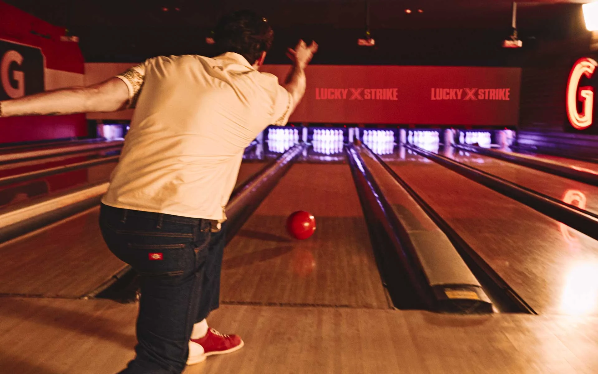 Man bowling at an upscale Lucky Strike venue, releasing a red bowling ball down polished lanes under warm ambient lighting with glowing pins in the distance.