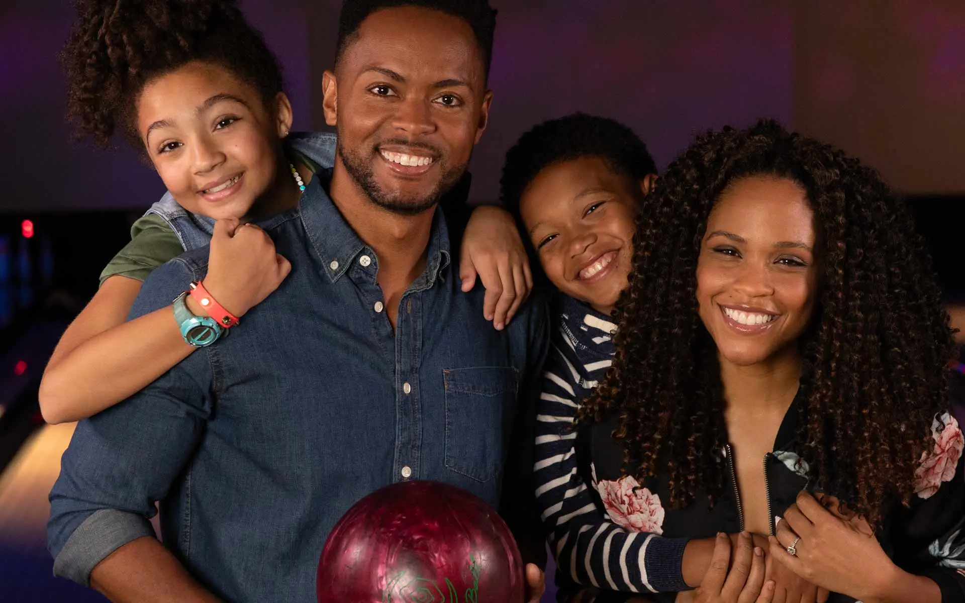 Smiling family at a bowling alley with two kids hugging their parents and a red bowling ball in the foreground.