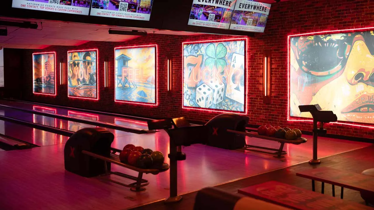 Bowling lanes at Lucky Strike Boca Raton lined with neon-framed murals reading 'Do You Feel Lucky' on an exposed brick wall with overhead screens.