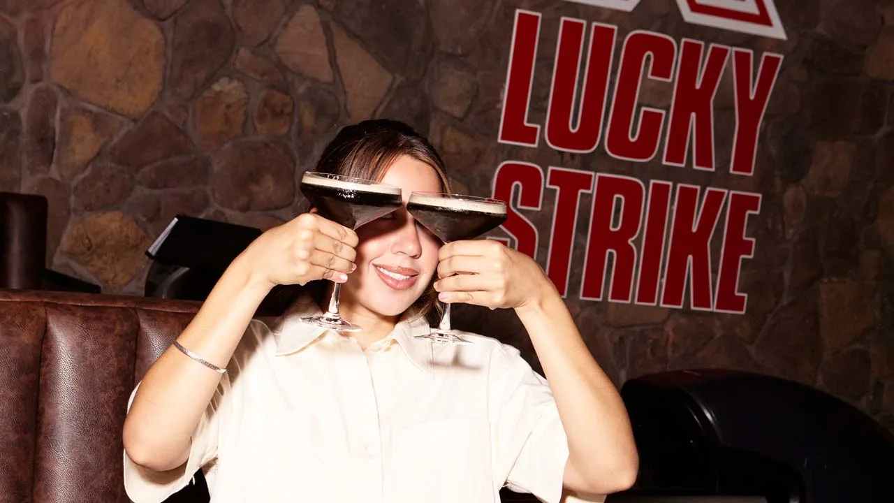 A woman holding two martinis in front of her face with a Lucky Strike sign in the background.