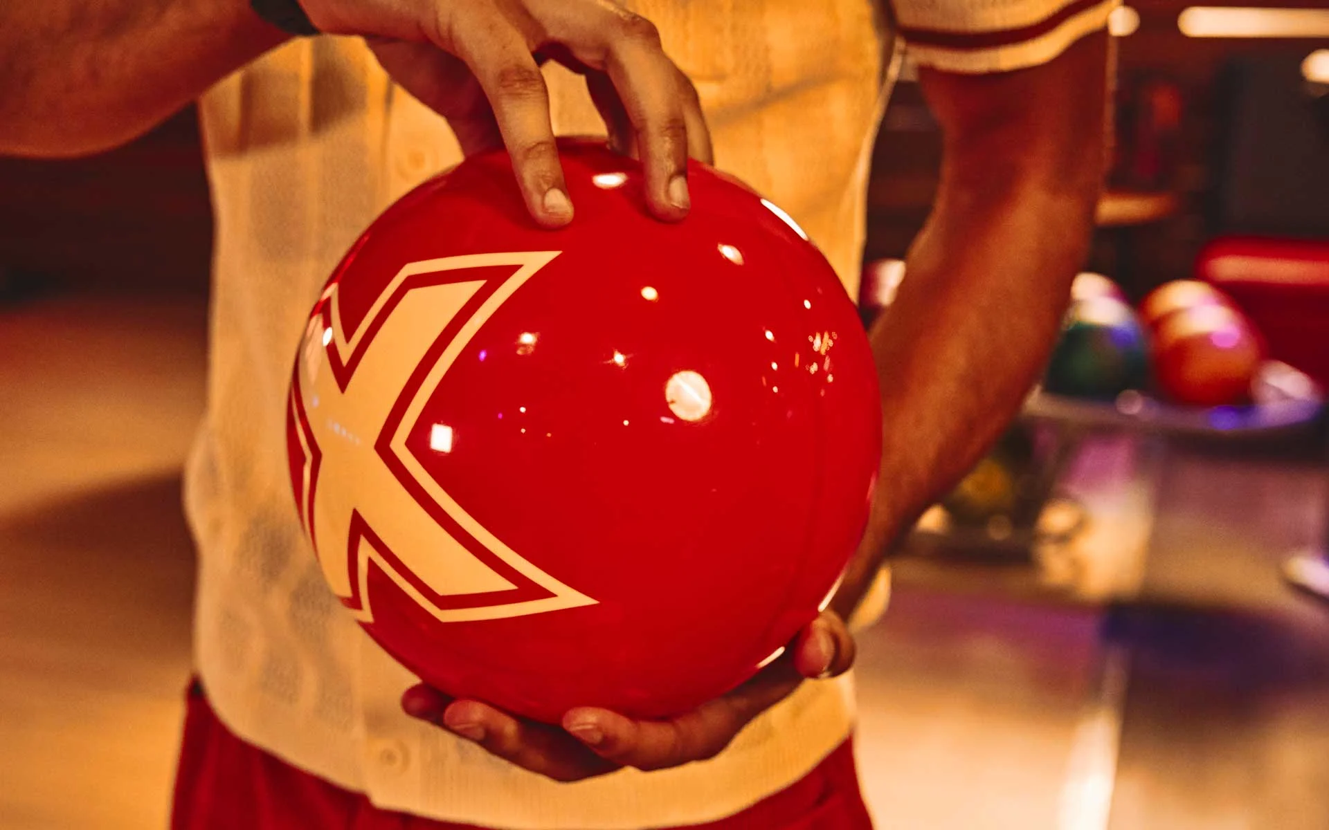 Close-up of hands holding a red bowling ball with a bold white “X” design in a warm-lit bowling alley setting.