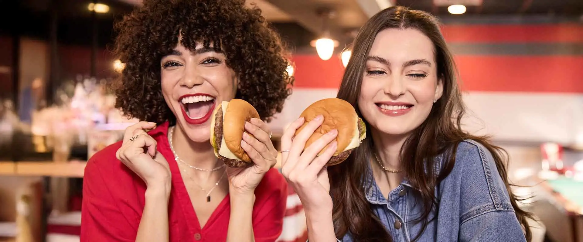 Two women laughing and holding burgers at AMF Bowling, enjoying food and drinks at the lanes.