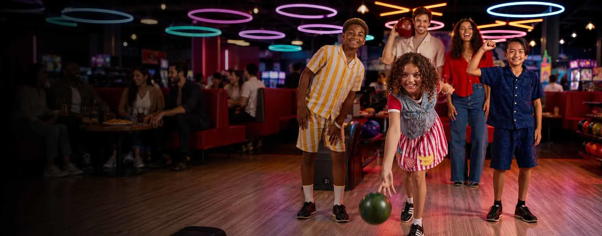 Families and friends bowling together at a vibrant bowling center with neon lighting, arcade games, and lounge seating.
