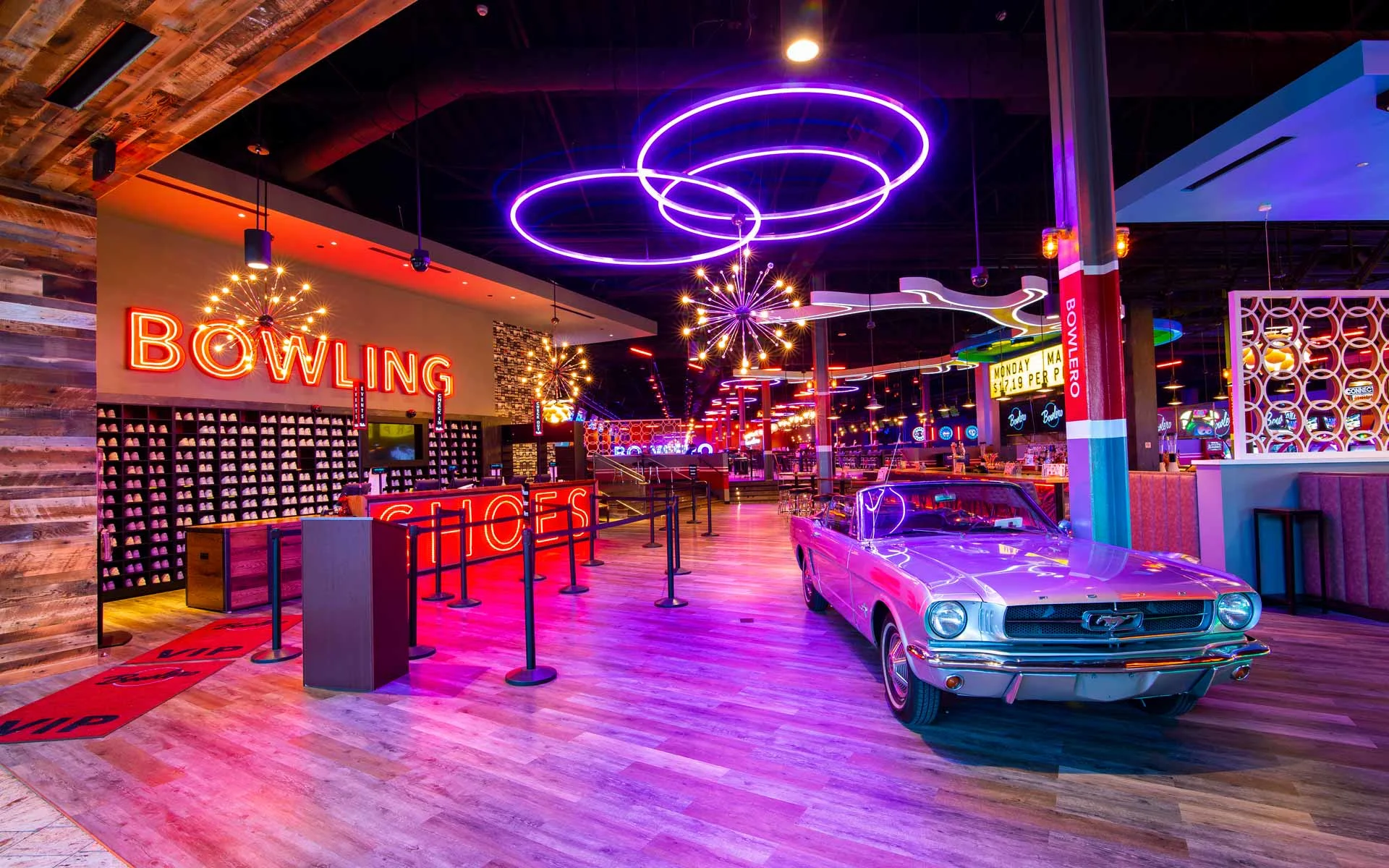 Upscale bowling center interior with vibrant neon lighting, featuring glowing circular ring lights overhead, a polished wood floor, and a retro silver convertible displayed near the entrance. To the left, a shoe rental area is lined with shelves of bowling shoes and illuminated signage, while the space opens into a lively, modern entertainment venue with warm ambient lighting, bold colors, and a stylish, nightlife-inspired atmosphere.