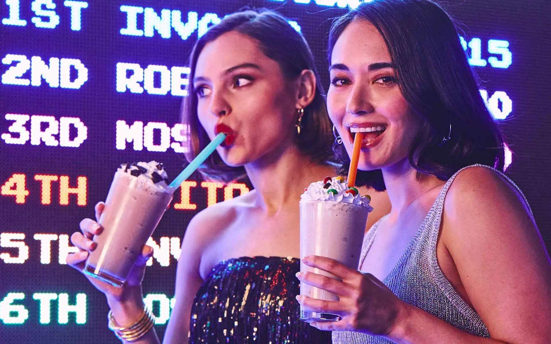 Two women sipping milkshakes in front of a glowing arcade high score screen.