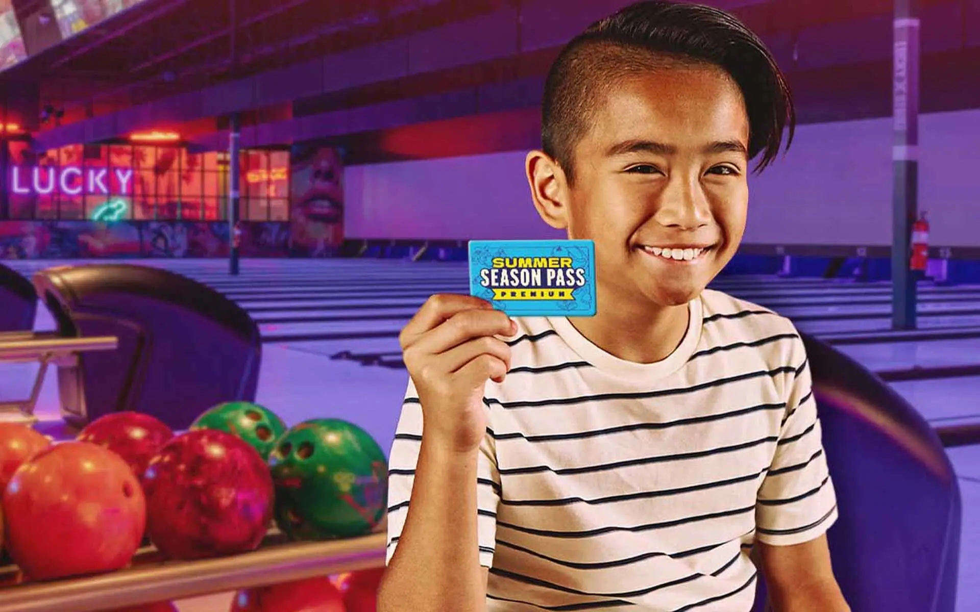 Smiling child holding a Lucky Strike Summer Season Pass inside a bowling alley with colorful bowling balls and lanes in the background.