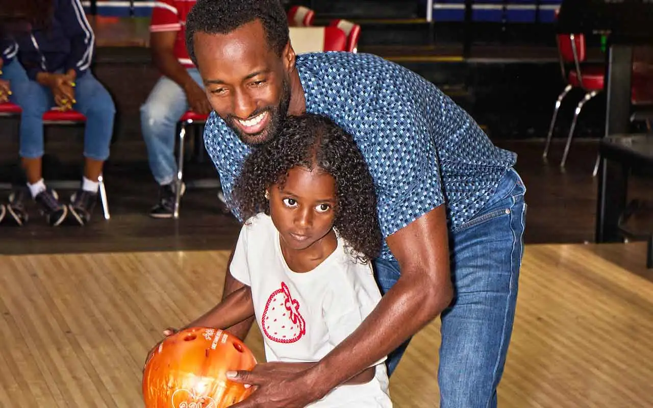Father helping his young daughter hold an orange bowling ball on the lanes at an AMF bowling center.