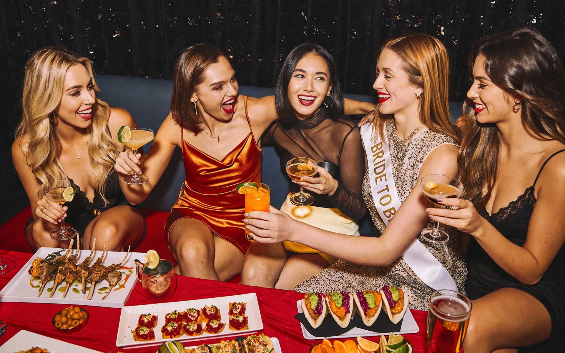 Five women at a bachelorette party toast cocktails in a lounge booth, with the bride-to-be wearing a sash on the right and appetizers spread across the table.