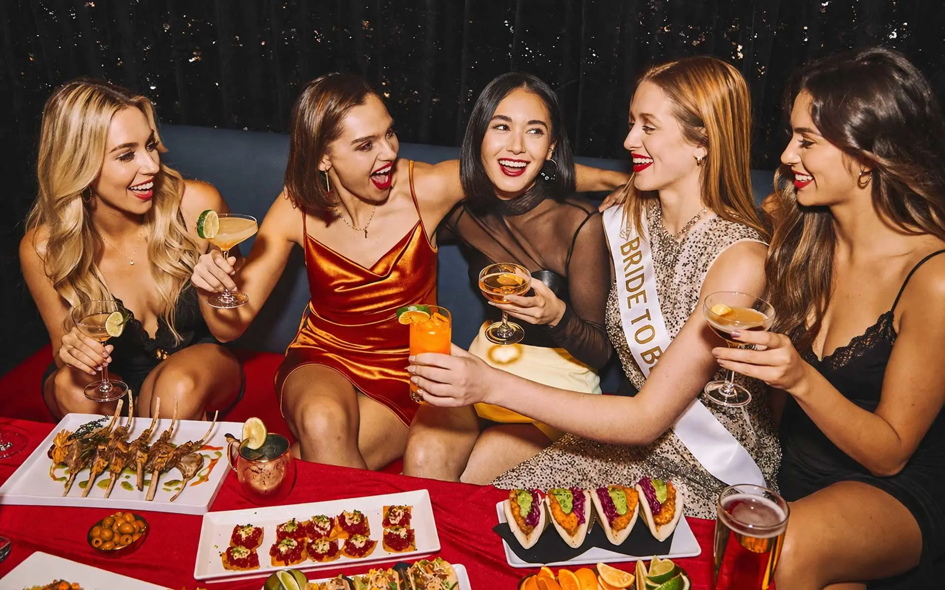 Five women at a bachelorette party toast cocktails in a lounge booth, with the bride-to-be wearing a sash on the right and appetizers spread across the table.