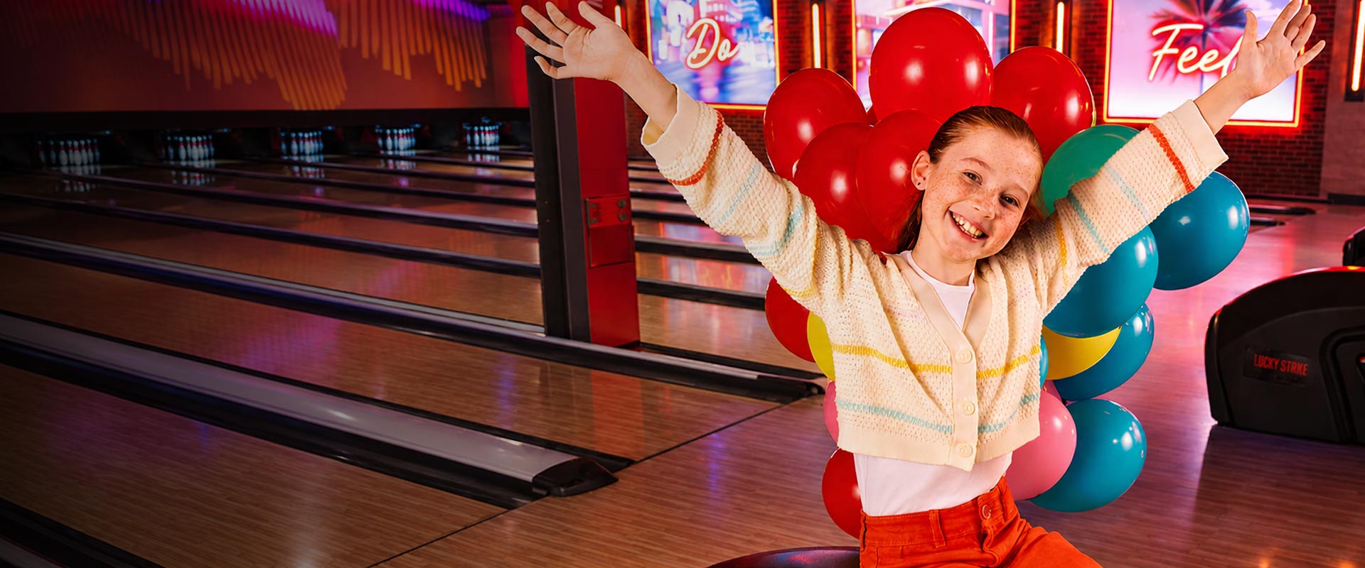 Smiling young girl sitting on a bowling ball return with arms raised, surrounded by colorful balloons at a Lucky Strike bowling center.