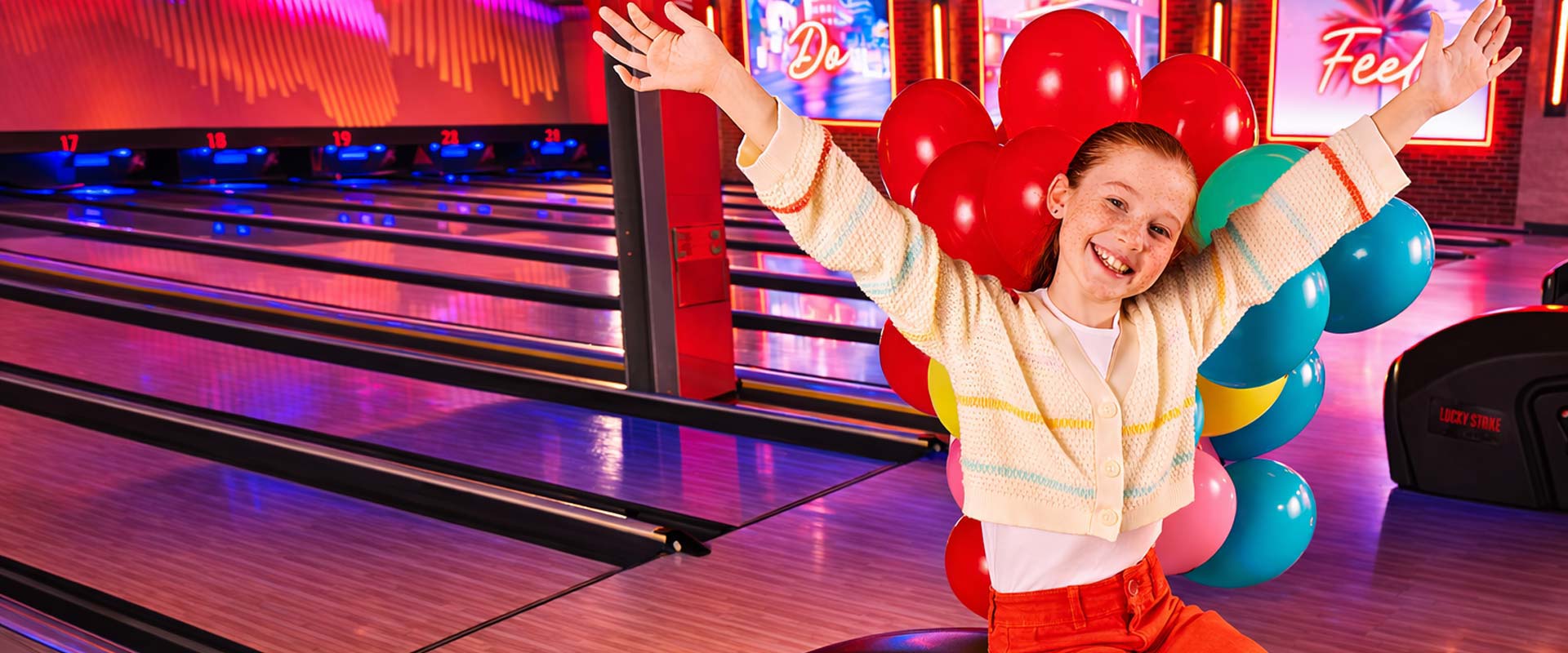 Smiling young girl sitting on a bowling ball return with arms raised, surrounded by colorful balloons at a Lucky Strike bowling center.