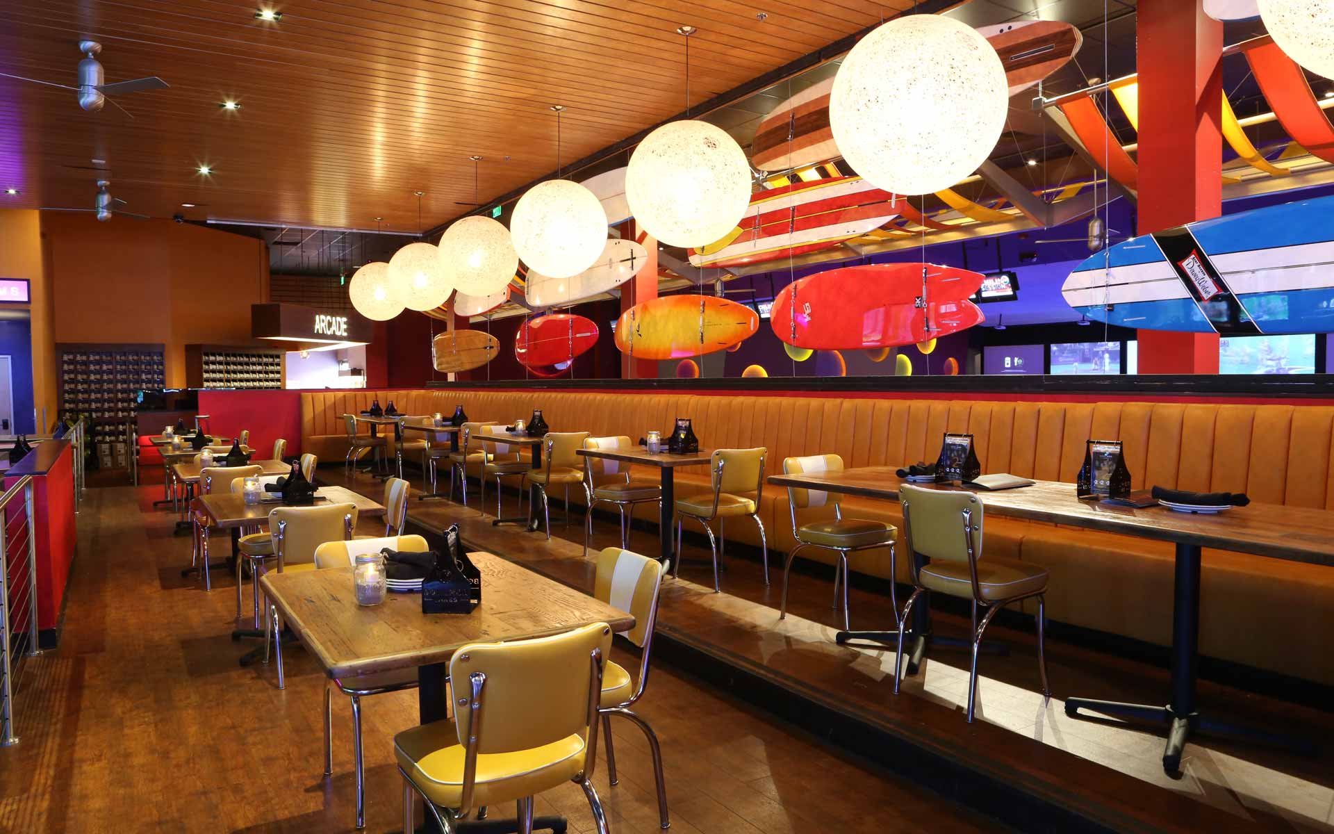 Upscale bowling lounge seating area with colorful surfboard decor, round pendant lighting, and lane-side tables at a modern entertainment venue.