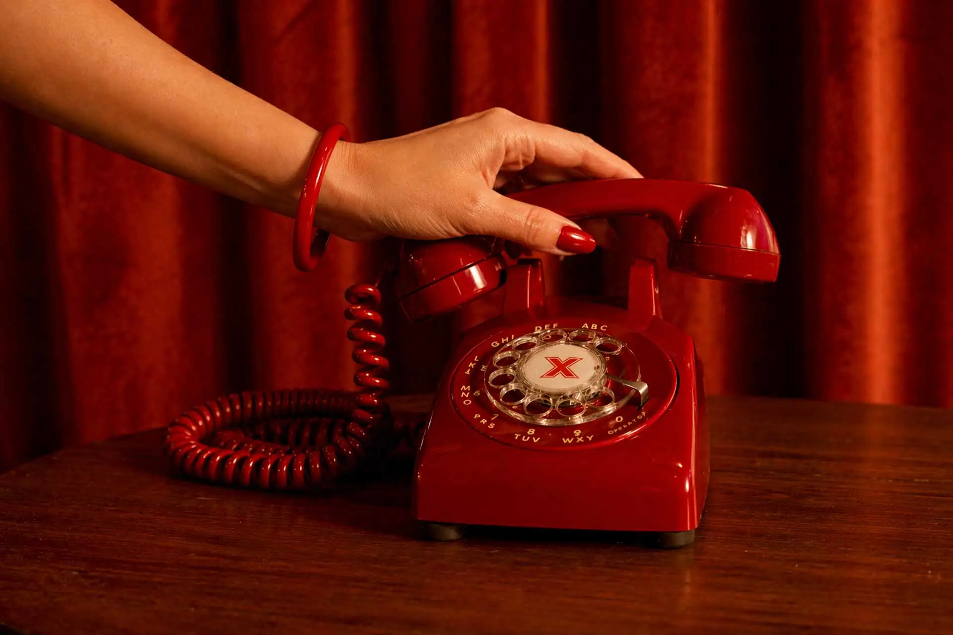 Close-up of a woman’s hand with red nail polish lifting the receiver of a red rotary phone on a wooden table, set against rich red velvet curtains.