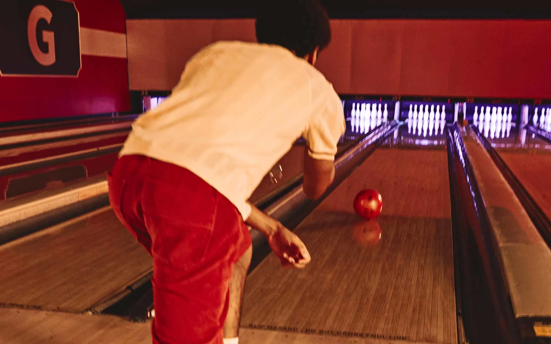 Person in red shorts releasing a bowling ball down a lane, viewed from behind, with pins illuminated at the end of the alley