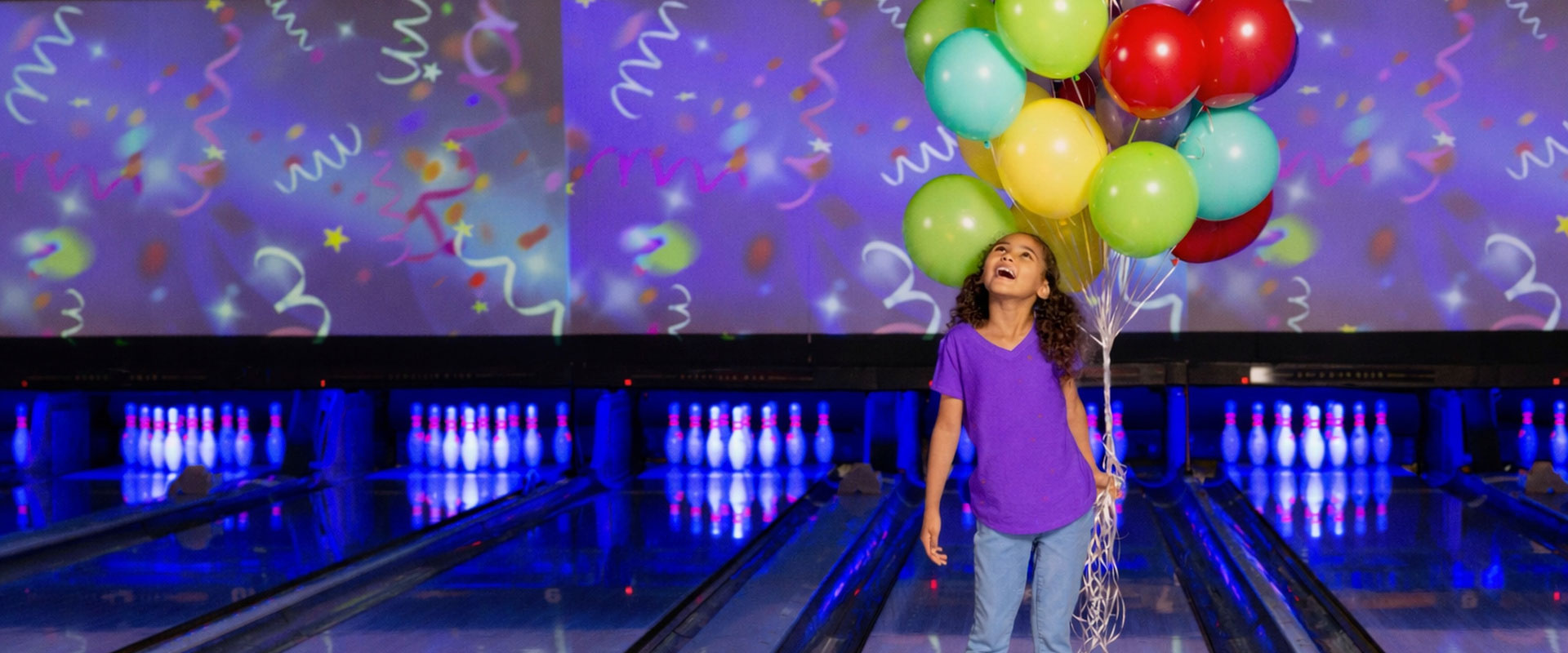 Young girl holding colorful balloons stands on a bowling lane in a neon-lit bowling alley with festive party graphics on the screens behind her.