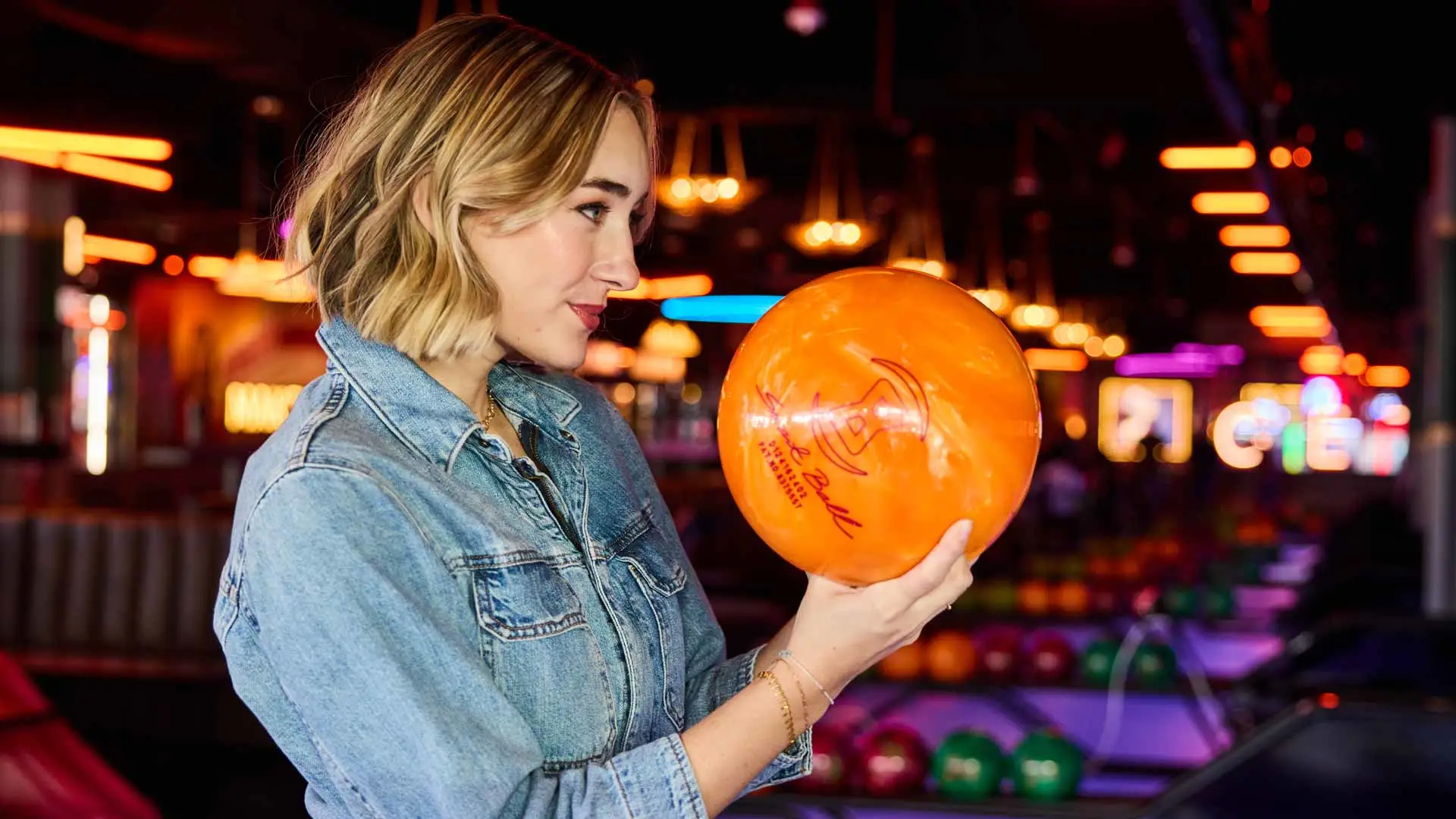 Woman holding an orange bowling ball inside a modern bowling alley with colorful lane lighting in the background.
