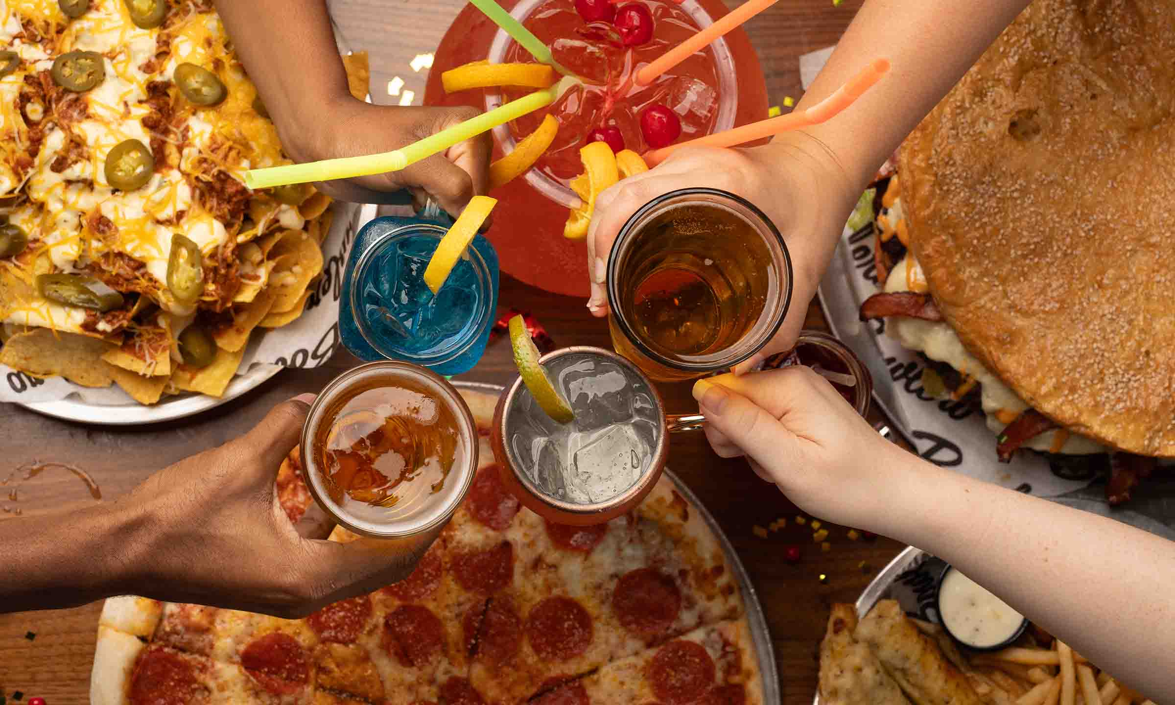 Four drinks coming together in a group cheers over pizza and nachos and a large sharable drink