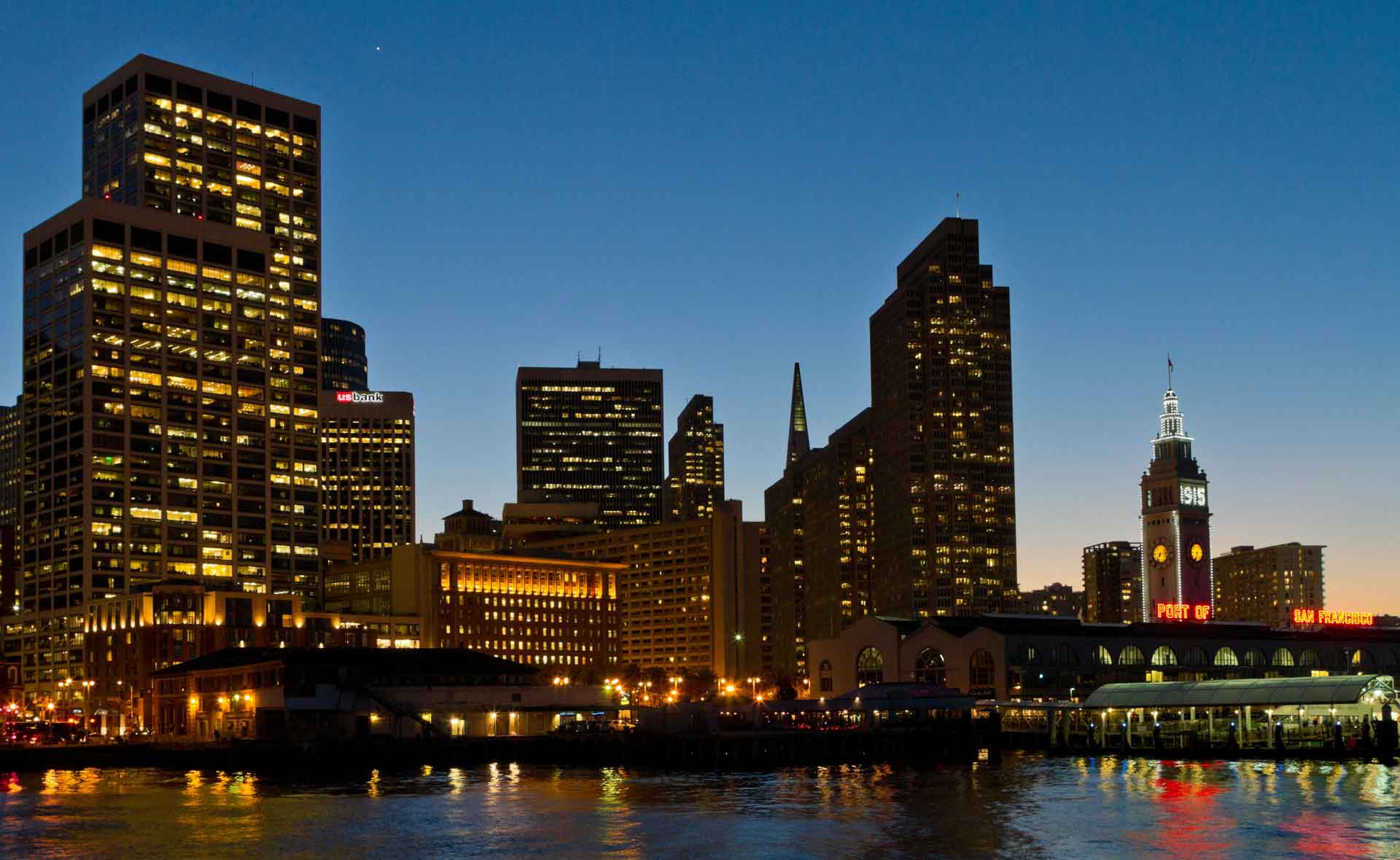 San Francisco skyline at dusk with illuminated downtown buildings and waterfront reflections along the bay.