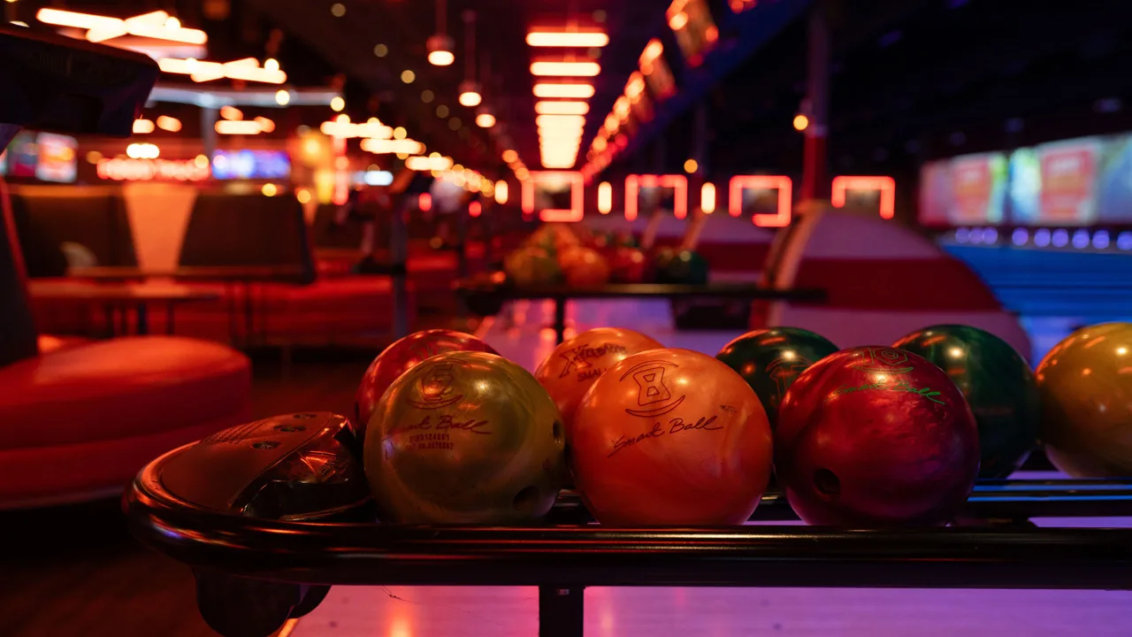 Colorful bowling balls in a return rack with bowling lanes and large TV screens in the background.