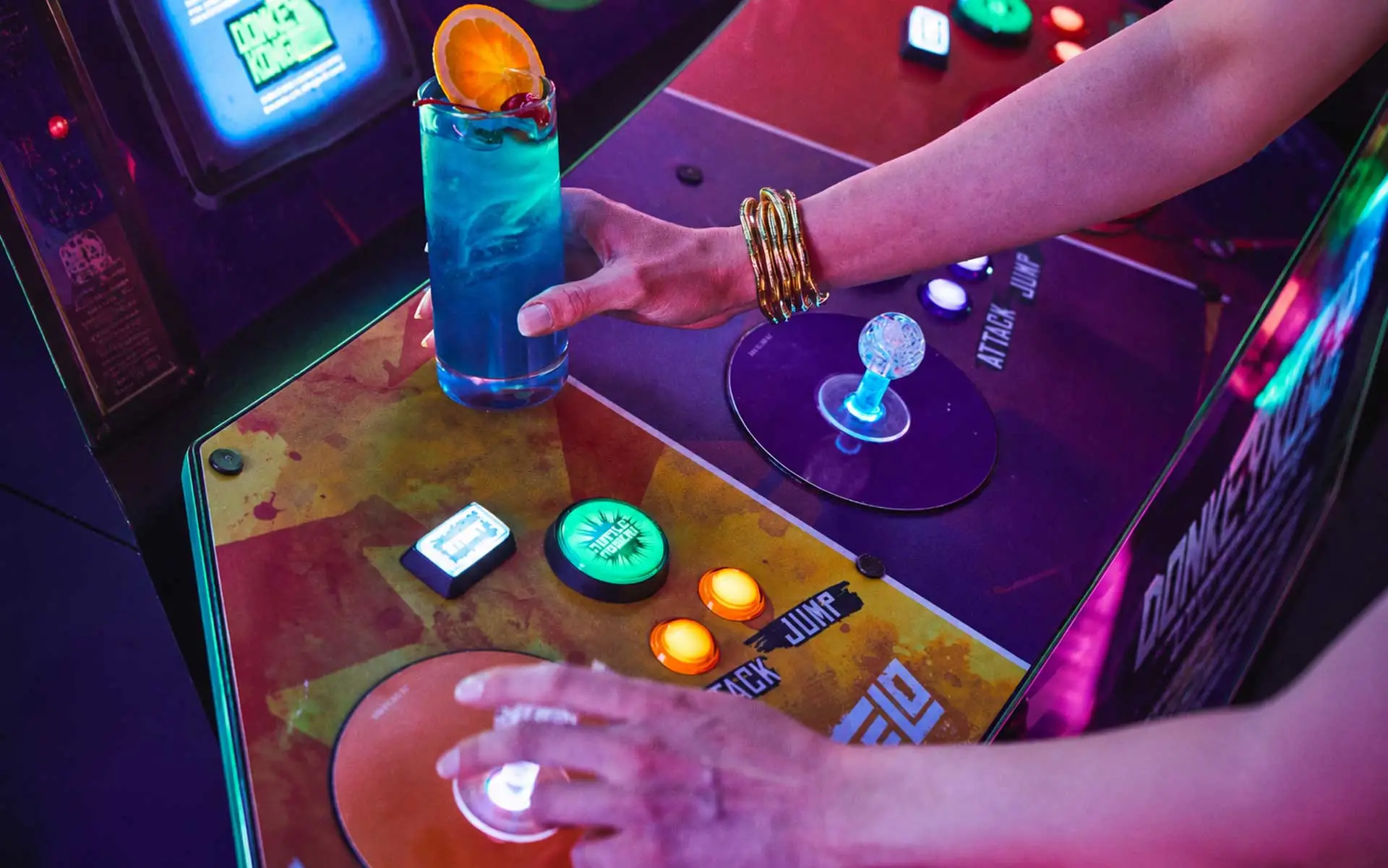 Woman holding a blue cocktail while playing an arcade game in a neon-lit entertainment venue with colorful glowing controls.