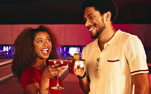 Two people smiling and holding cocktails with bowling lanes in the background