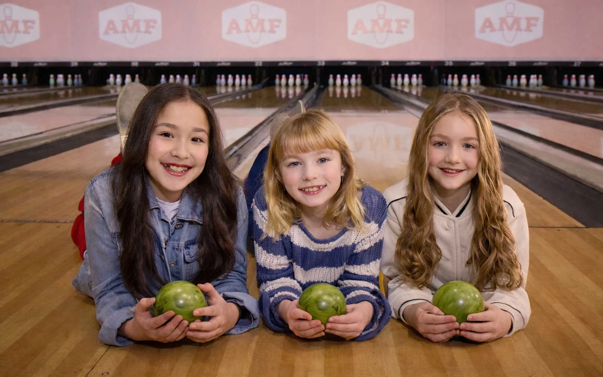 Three smiling children lie on a polished AMF bowling lane, each holding a small green duckpin bowling ball with no finger holes, while duckpin pins are set at the end of the lanes in the background.