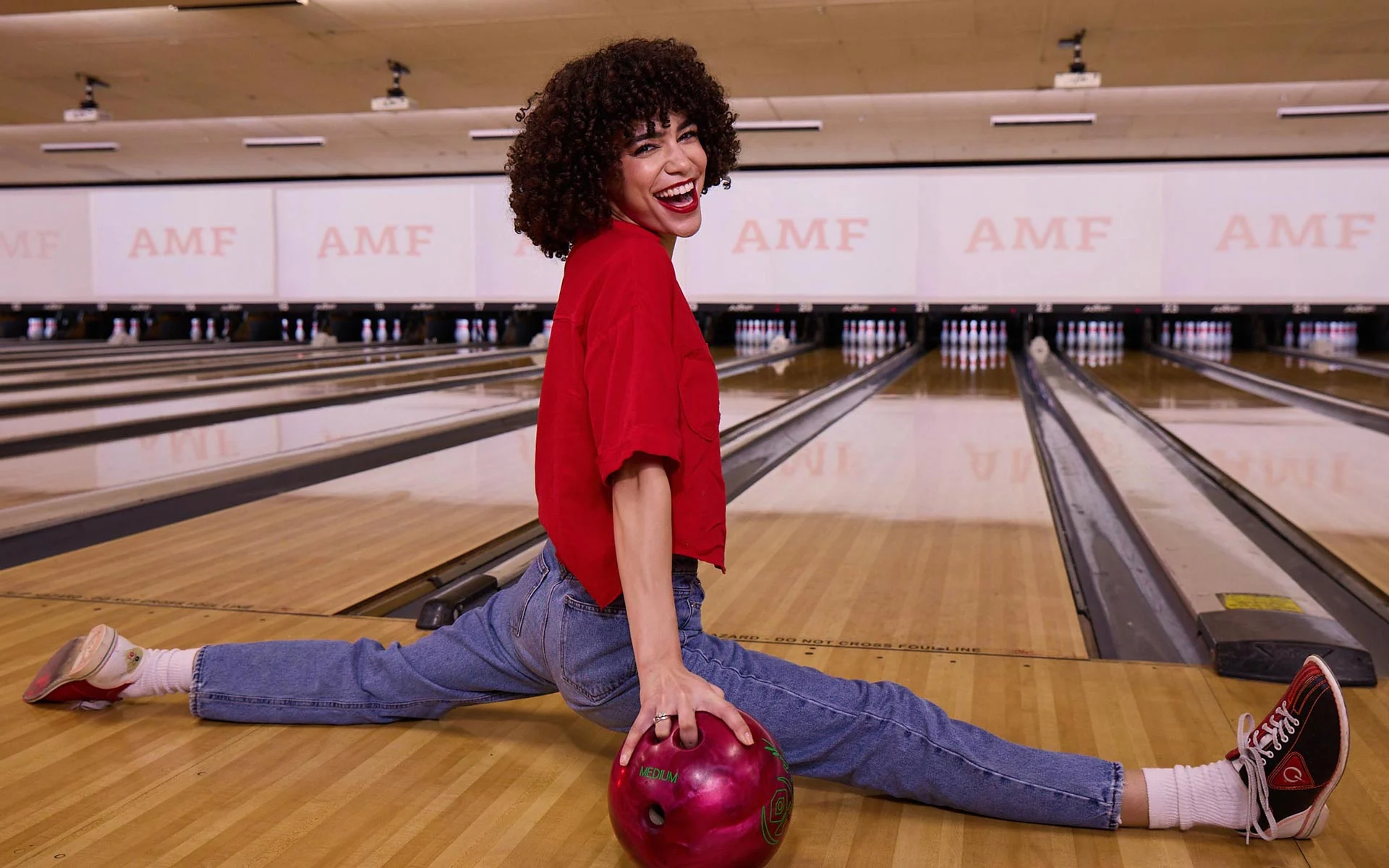 Smiling woman doing a split on a bowling lane while holding a bowling ball at an AMF bowling center.