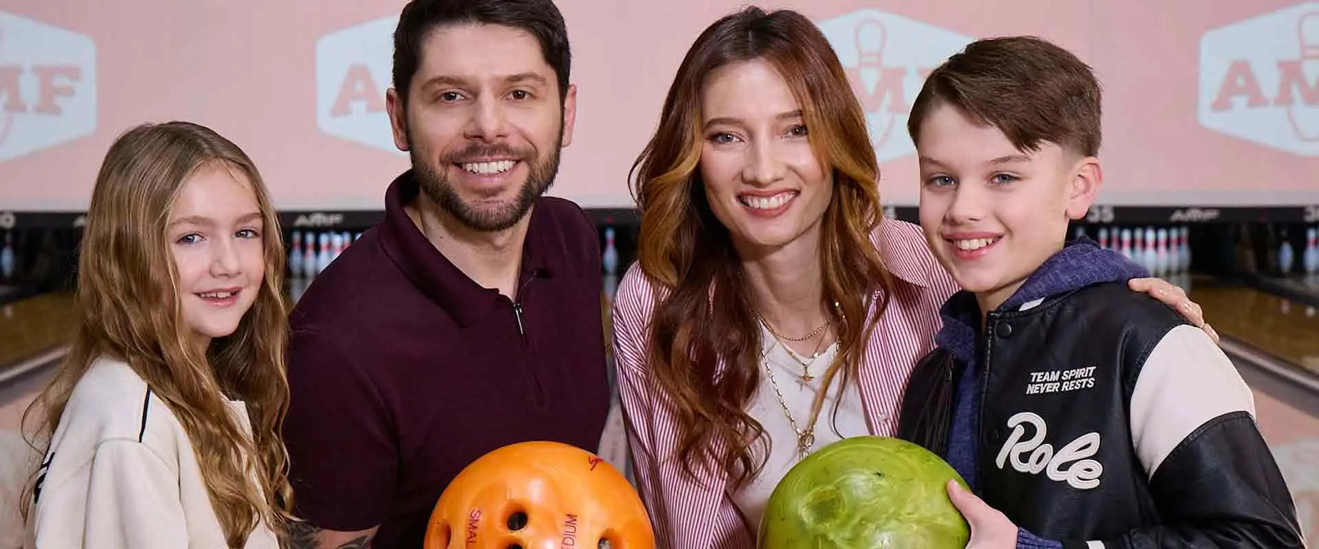 Family of four smiling and holding bowling balls at AMF Bowling center, ready to play.