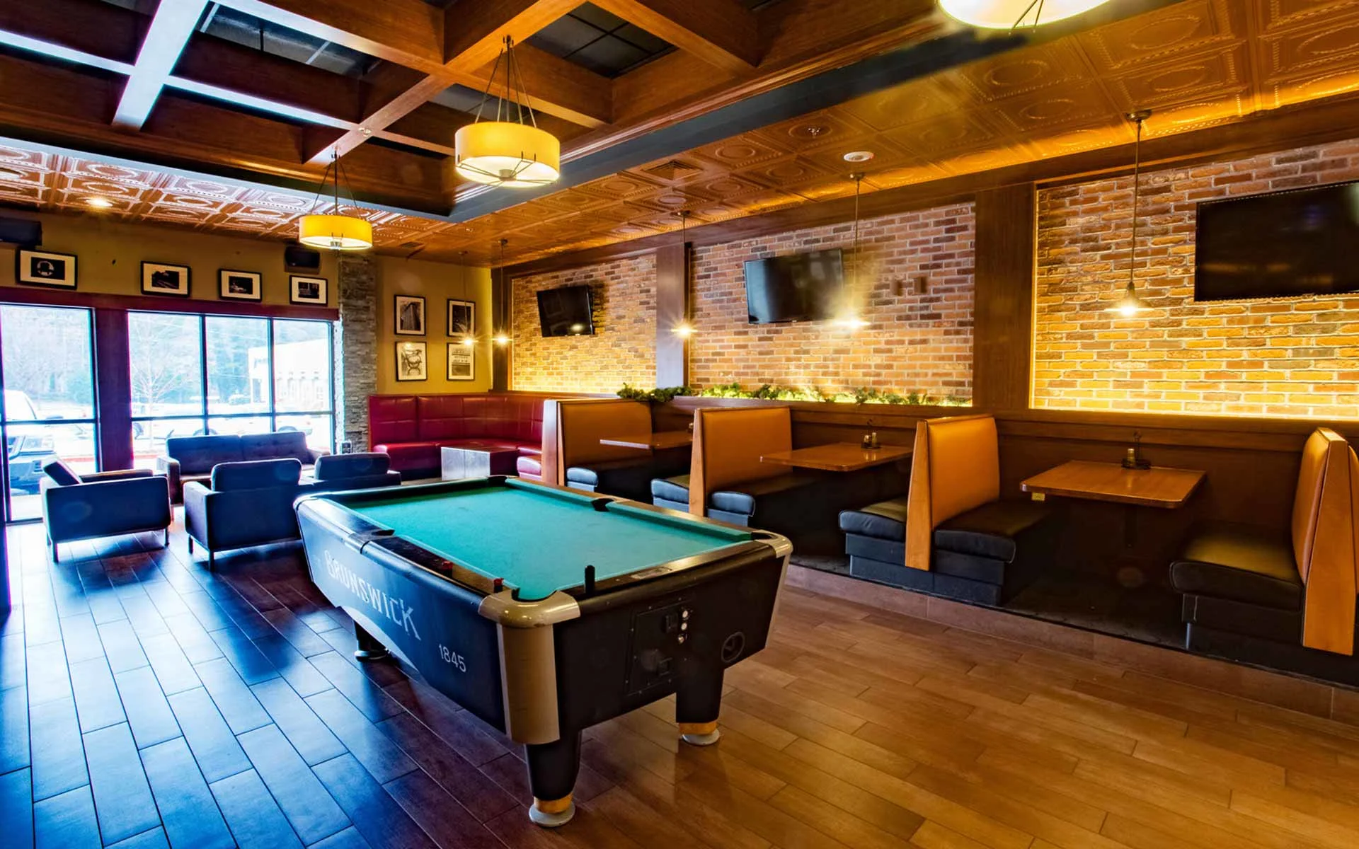 Upscale lounge area with a pool table at center, surrounded by cozy booth seating, brick accent walls, warm pendant lighting, and mounted TVs, creating a relaxed and stylish social space