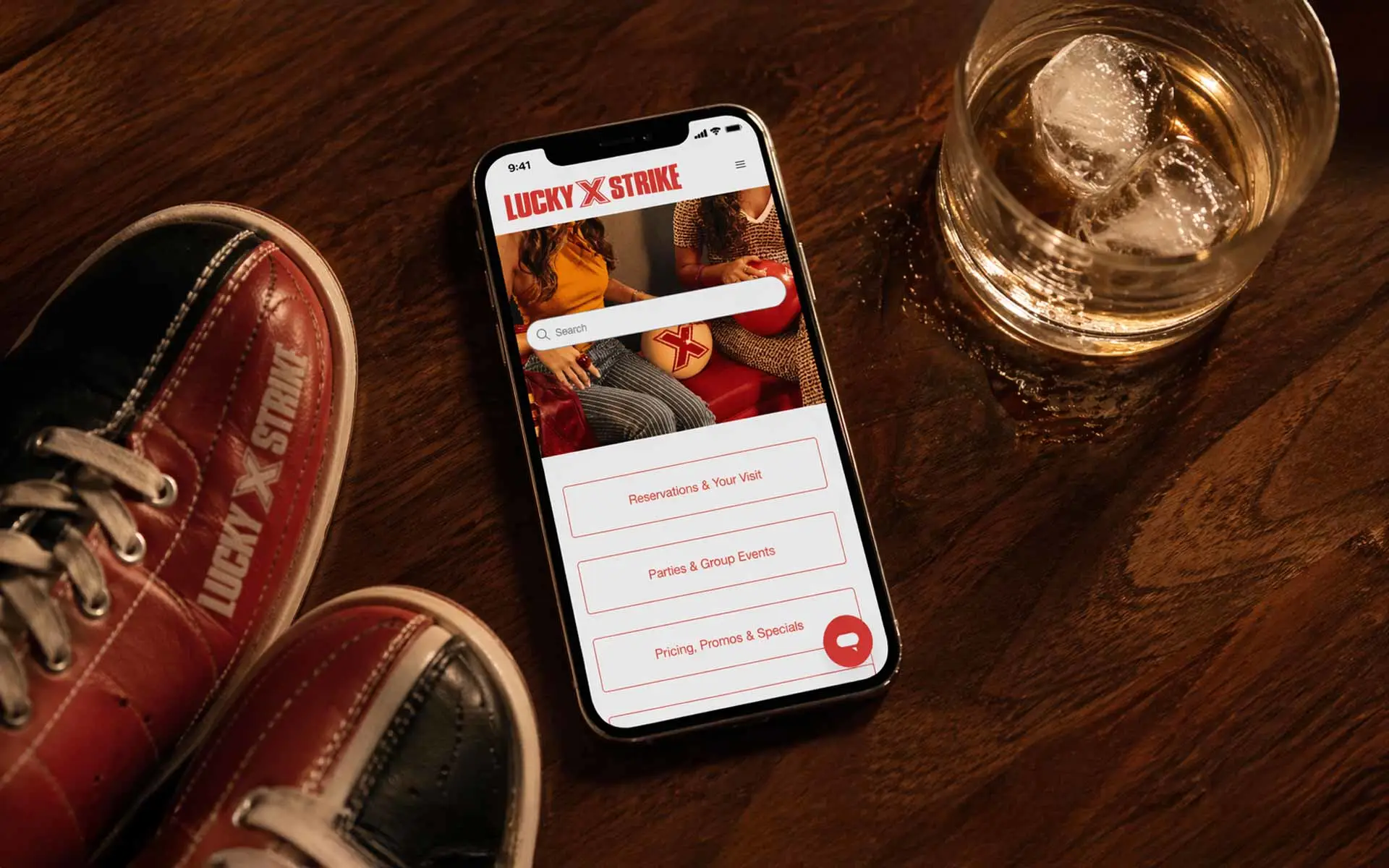 Overhead flat-lay of a smartphone on a walnut wood bar showing the Lucky Strike website, with a lowball cocktail glass and ice at the top right and red-and-white bowling shoes at the bottom left in warm ambient lighting.