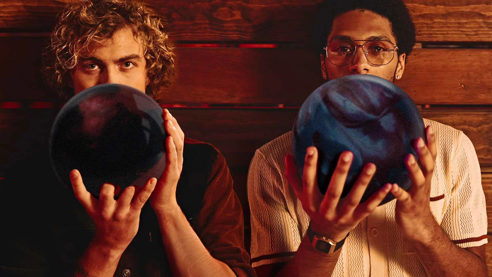 Two young men holding bowling balls in front of their faces, staring directly into the camera with a dramatic, warm-toned background.